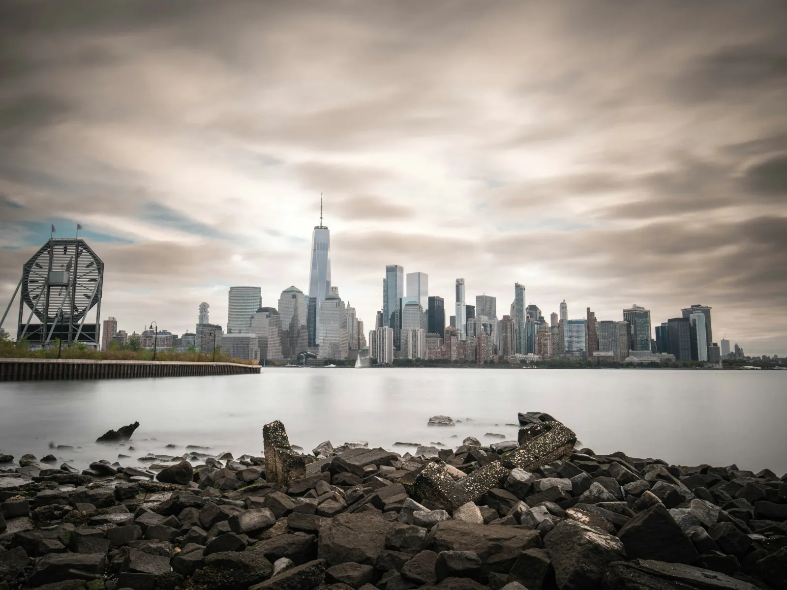 View of New York City skyline from rocky shore with cloudy sky and water in the foreground