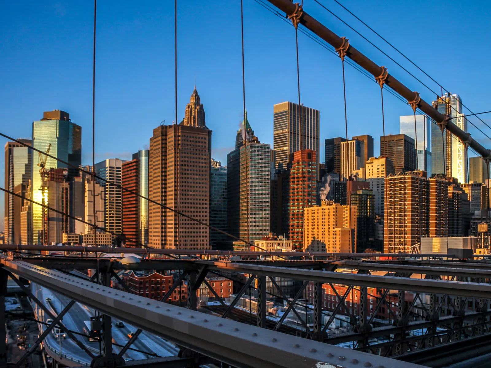 View of a city skyline with modern skyscrapers seen through the steel cables of a bridge during sunset.