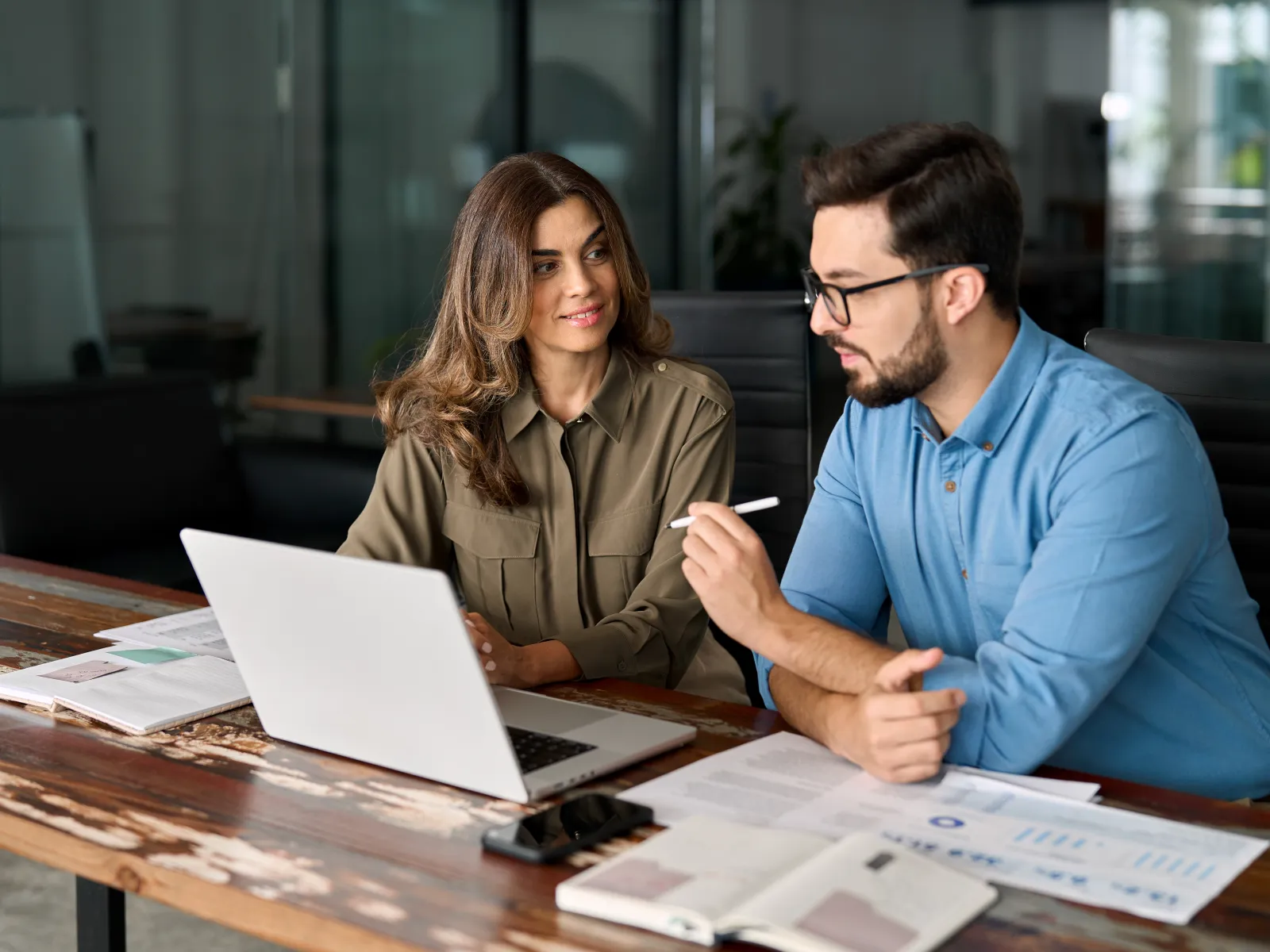 Two colleagues discussing work with laptop and documents in a modern office meeting room
