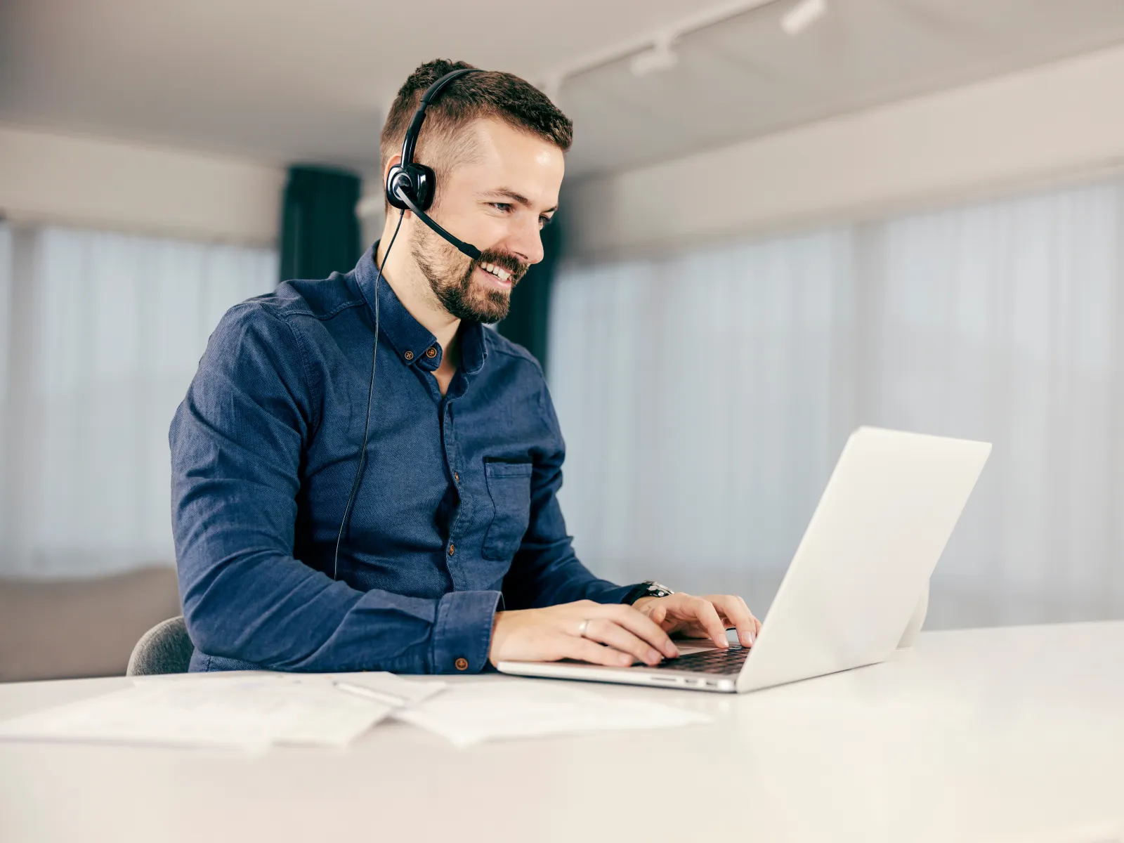 Man wearing headset working on laptop in bright office, smiling during online communication or support call