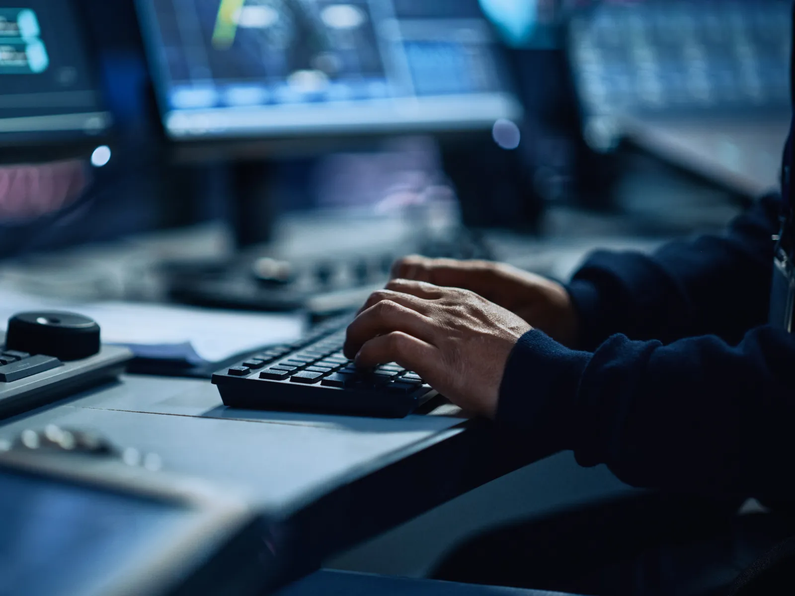 Person working on a computer keyboard with multiple monitors displaying data and graphs in a dark room.