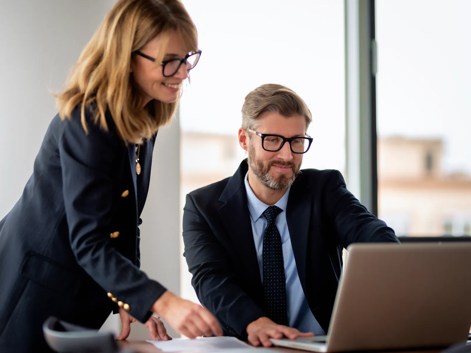 Two professionals in business attire collaborating at a desk with laptop and documents in an office setting