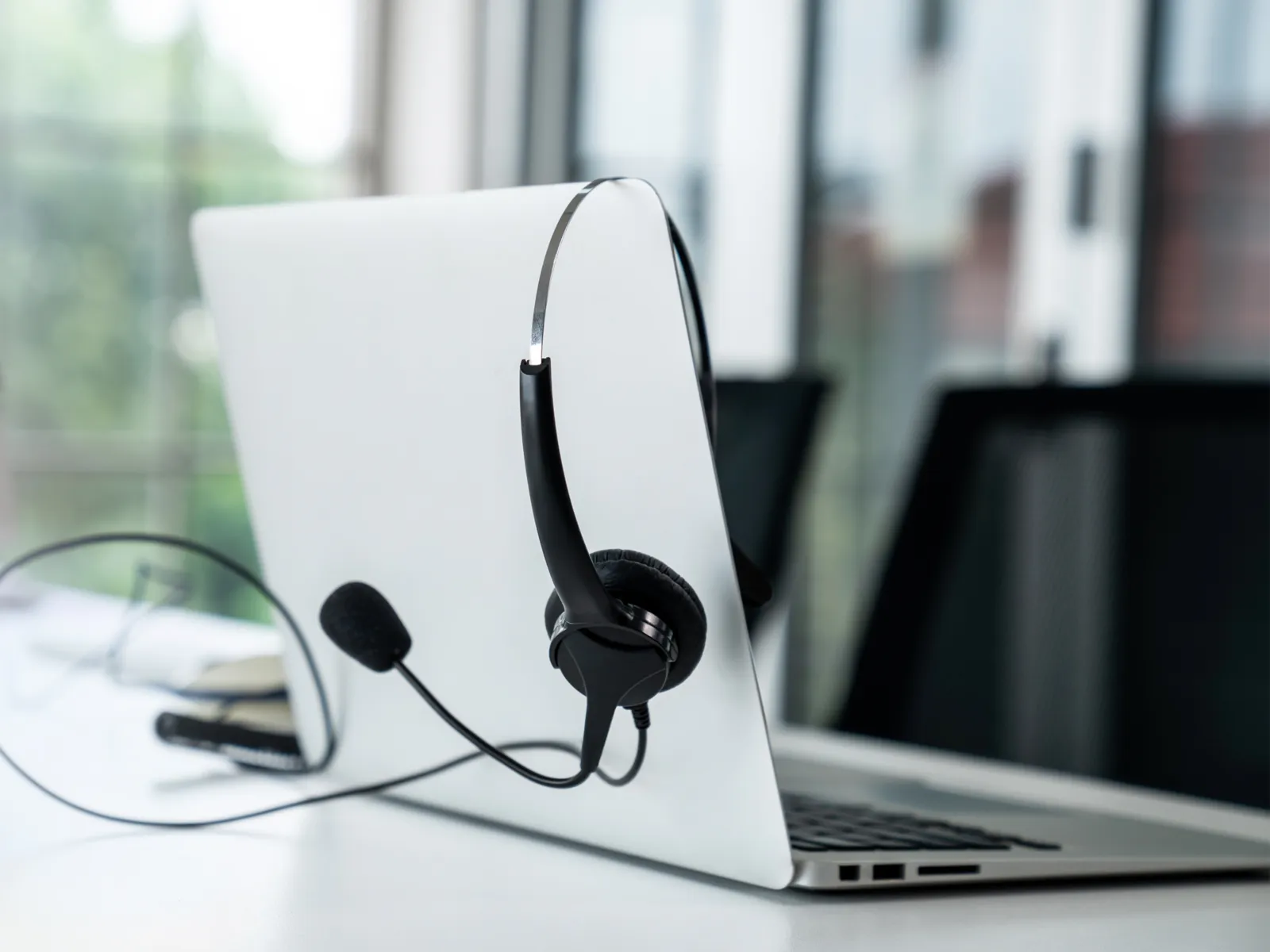 White laptop with headset resting on the screen on a clean white desk near a window with blurred background.