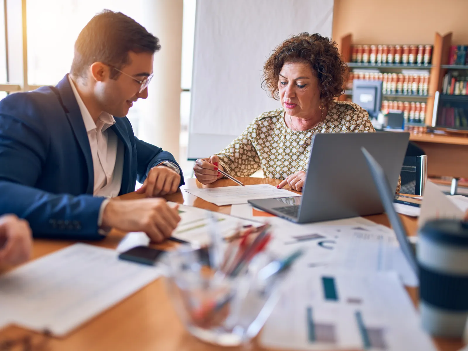 Business professionals collaborating over paperwork and laptop in a modern office with bookshelves.
