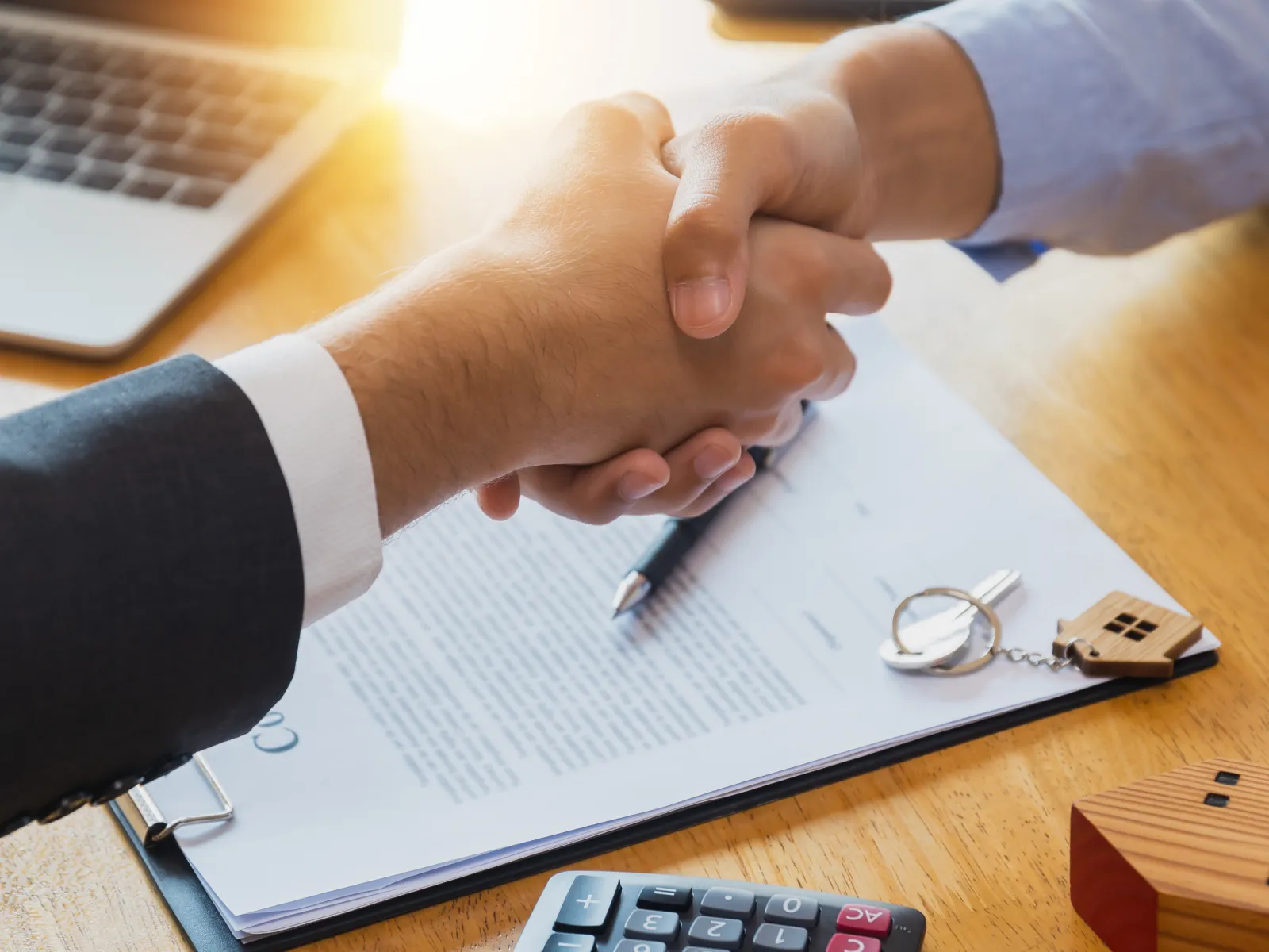Business handshake over signed contract with laptop, calculator, and house key on wooden desk
