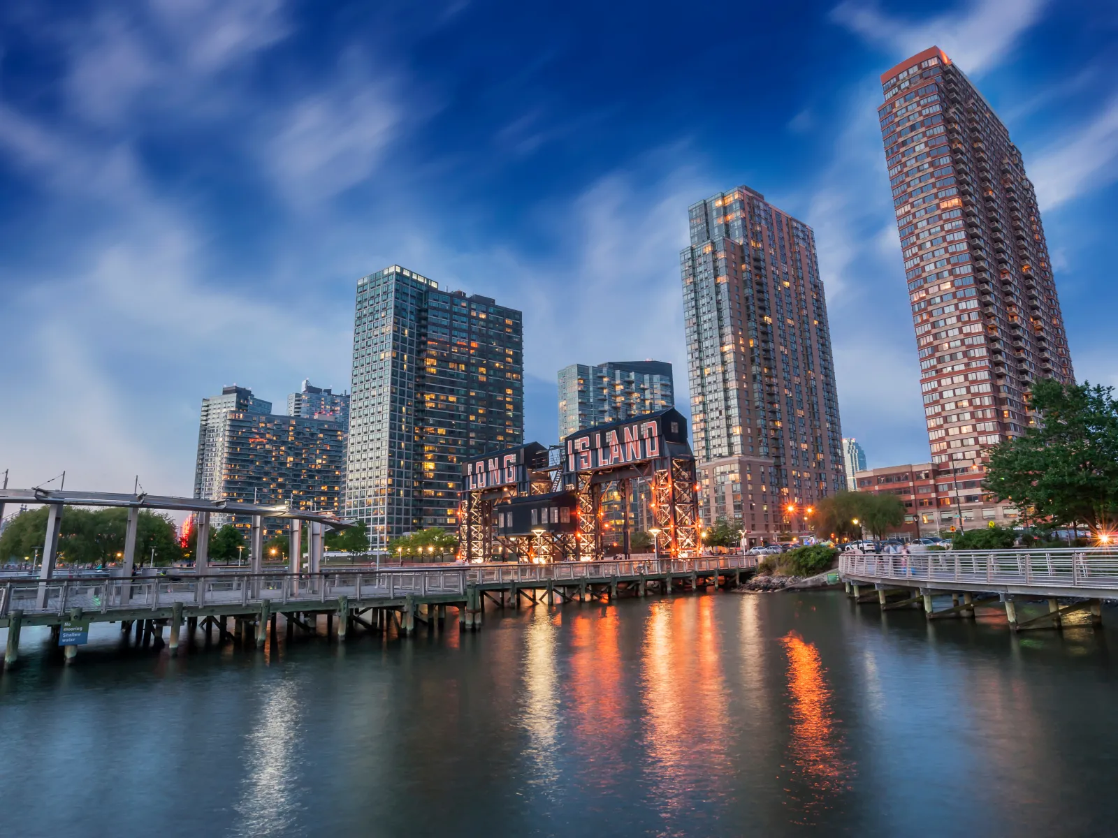 Long Island City waterfront with illuminated buildings and piers reflecting on water at dusk under a blue sky.