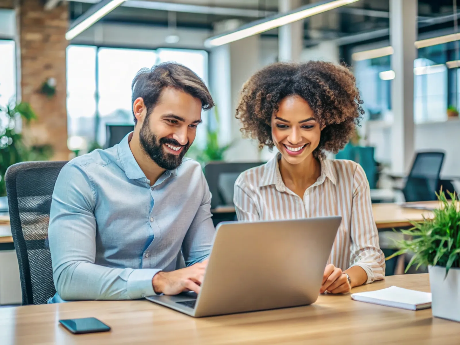 Two coworkers smiling and collaborating on a laptop in a modern bright office space with plants.