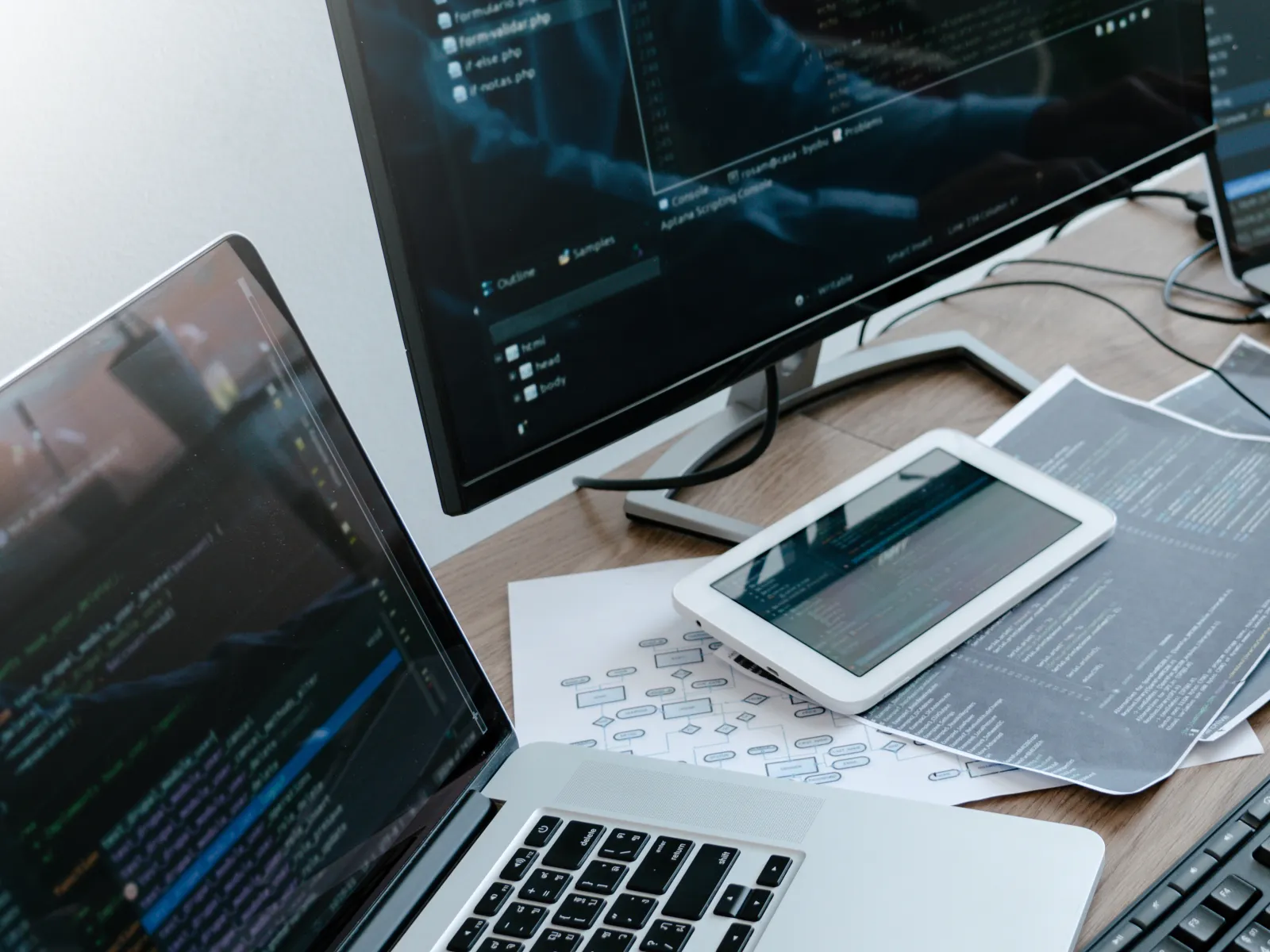 Person coding on laptop and dual monitors with tablets and technical papers on wooden desk
