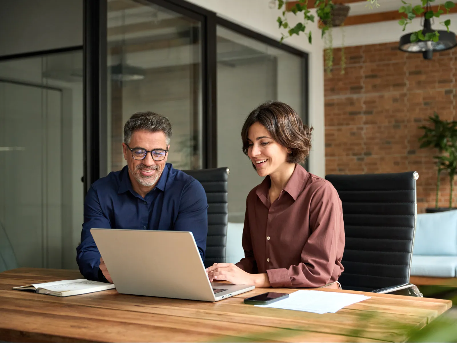 Two colleagues smiling and working together on a laptop in a modern office with brick walls and plants.