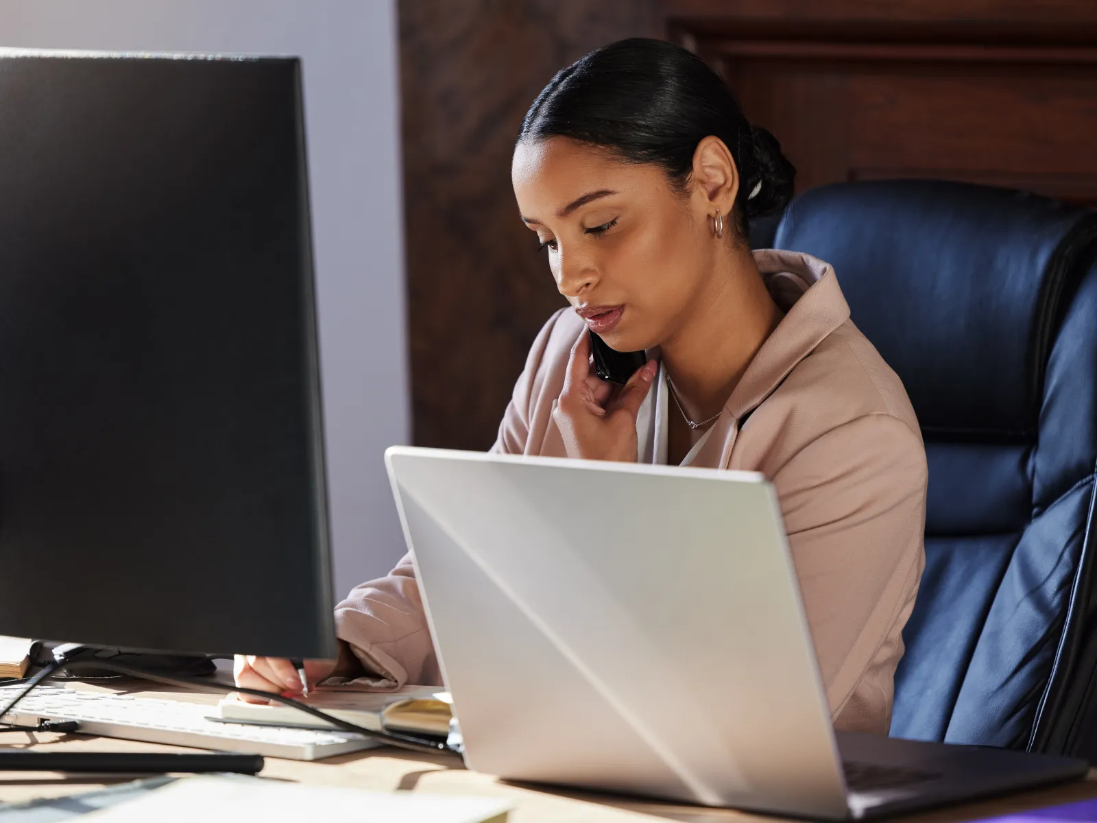 Professional woman multitasking with phone and computer while taking notes at office desk