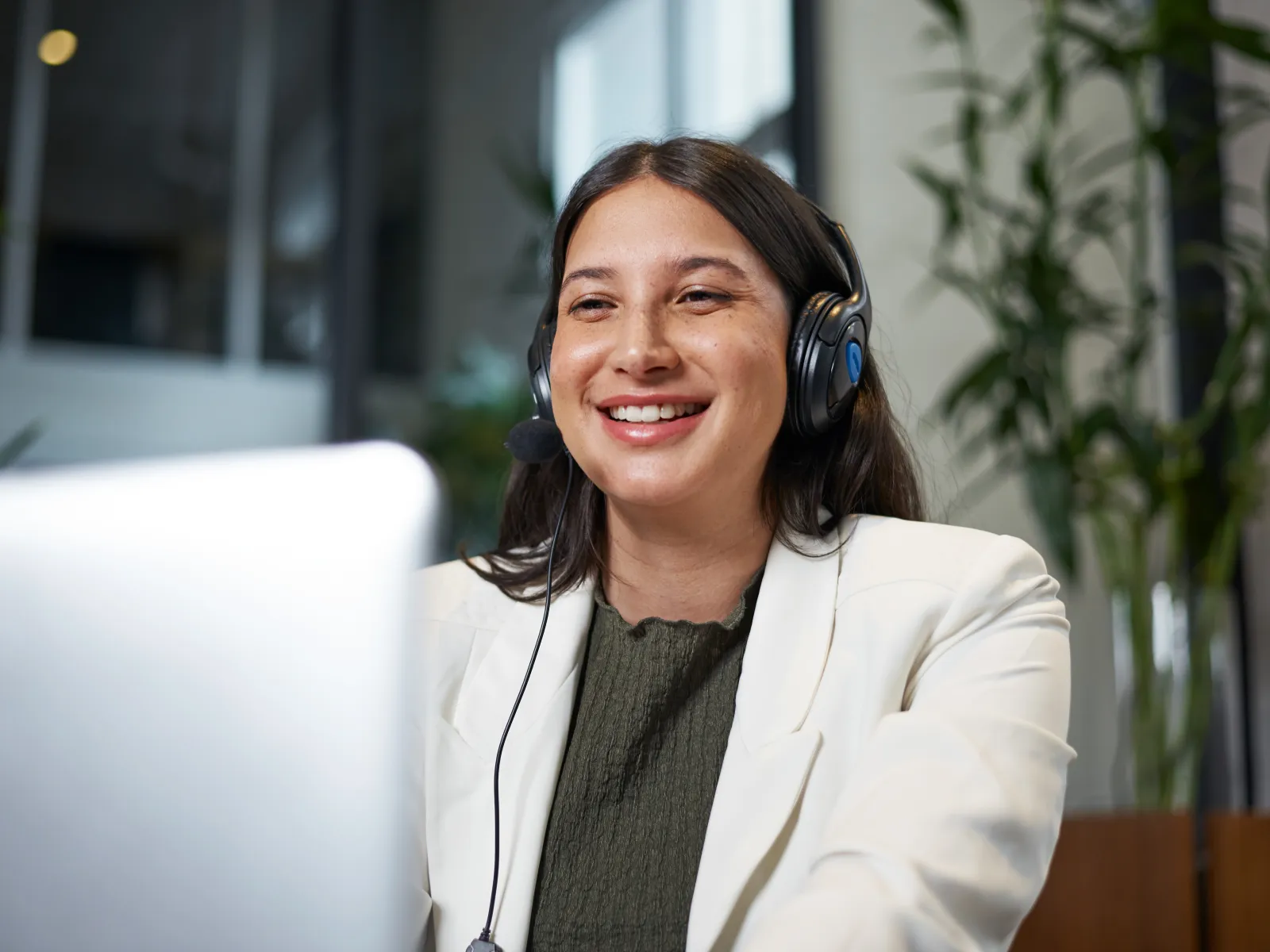 Smiling woman wearing headset and white blazer working on laptop in modern office environment