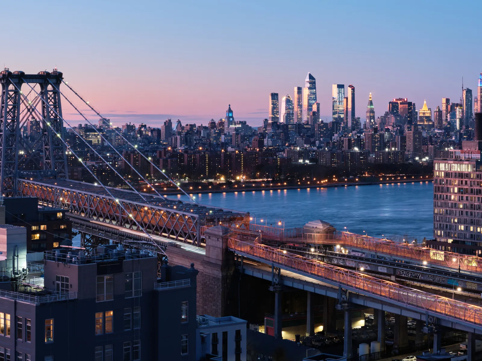 Sunset view of Williamsburg Bridge with illuminated New York City skyline and East River in the background.