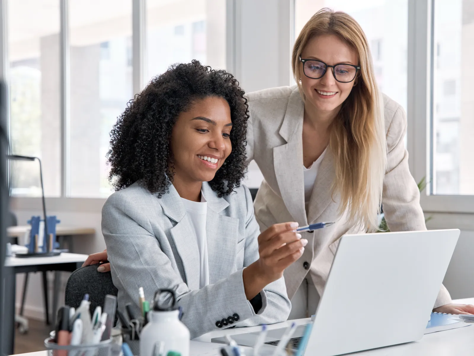 Two businesswomen collaborating in office, looking at laptop, discussing work with smiling expressions.