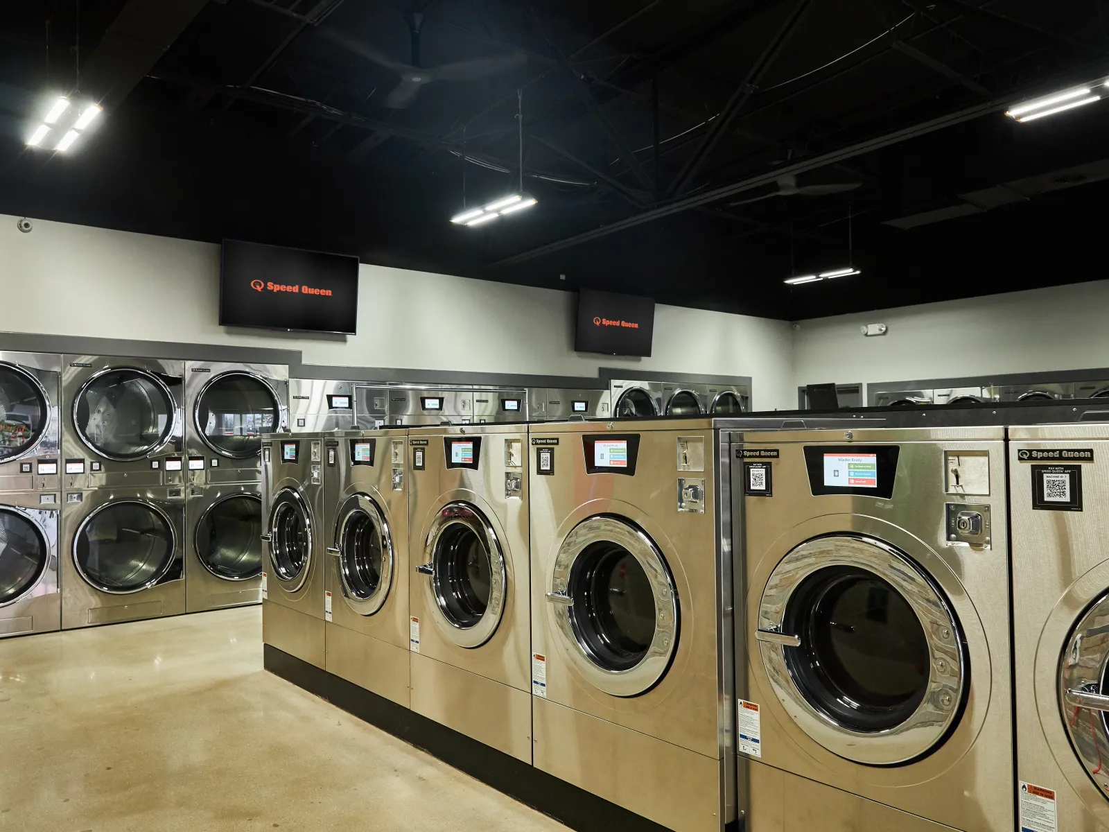 Interior of a modern laundromat with rows of stainless steel washing machines and dryers under bright LED lights.
