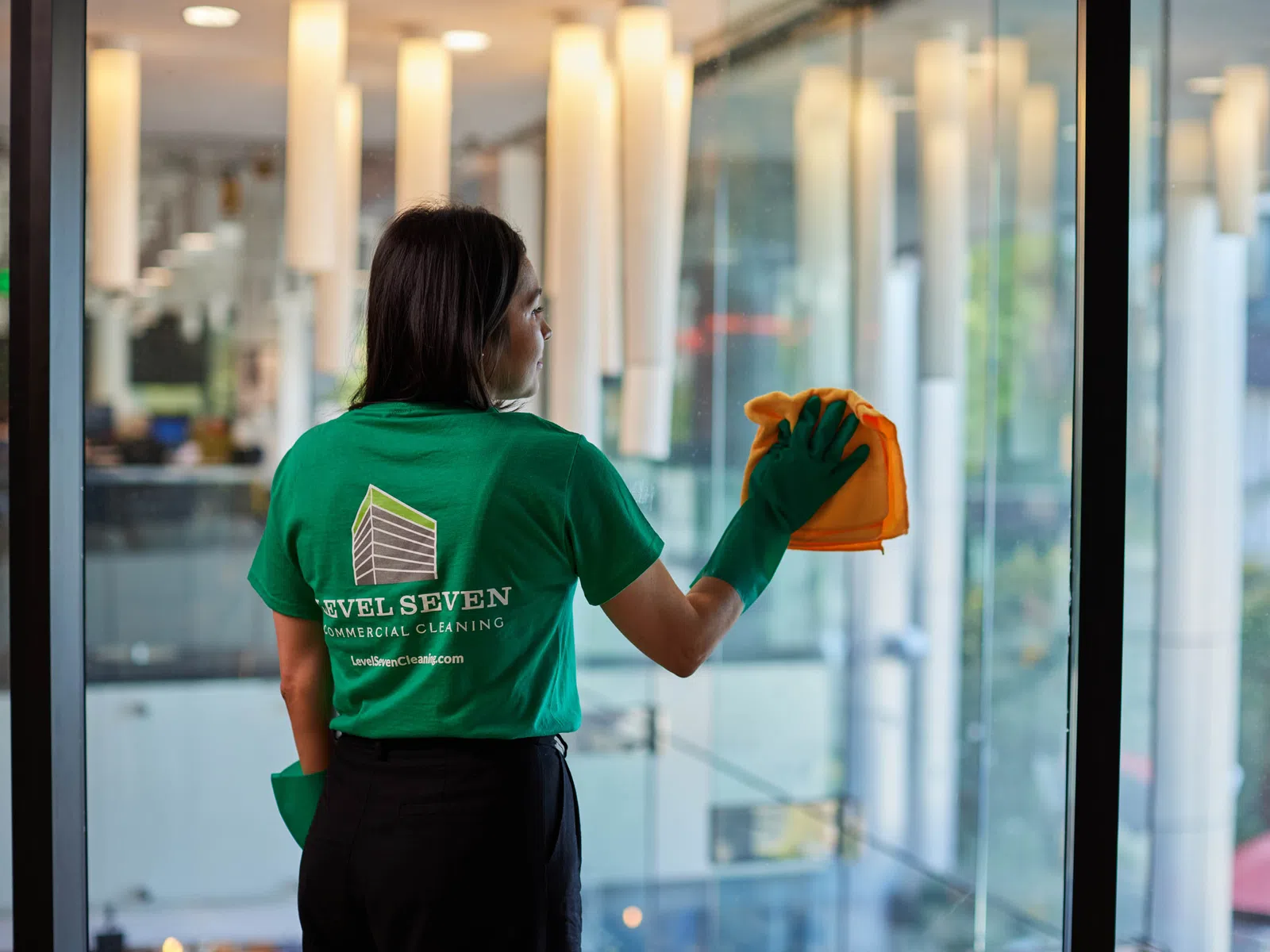 Woman wearing green Level Seven shirt cleaning glass window with a cloth inside a modern building.