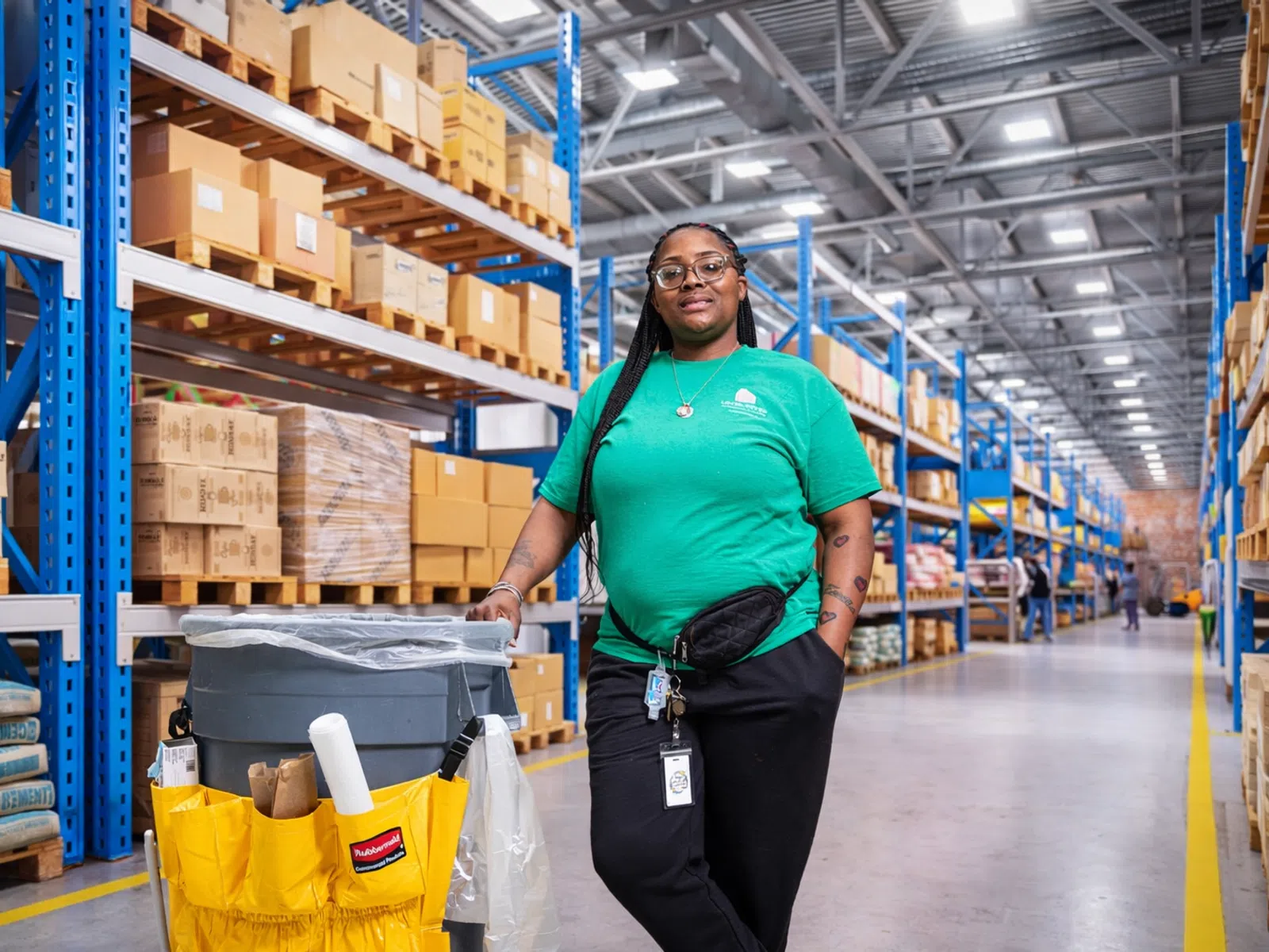 Warehouse worker in green shirt standing next to cleaning cart in aisle with shelves of boxes and supplies.