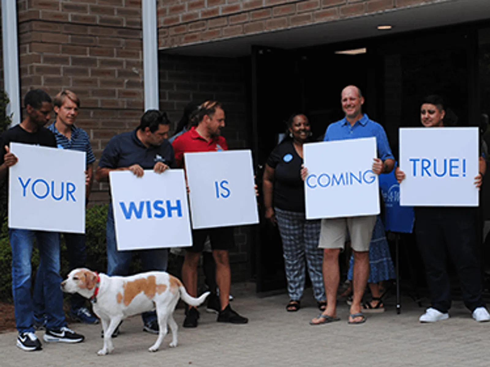Group of people holding signs reading Your wish is coming true with a dog in front of a brick building.