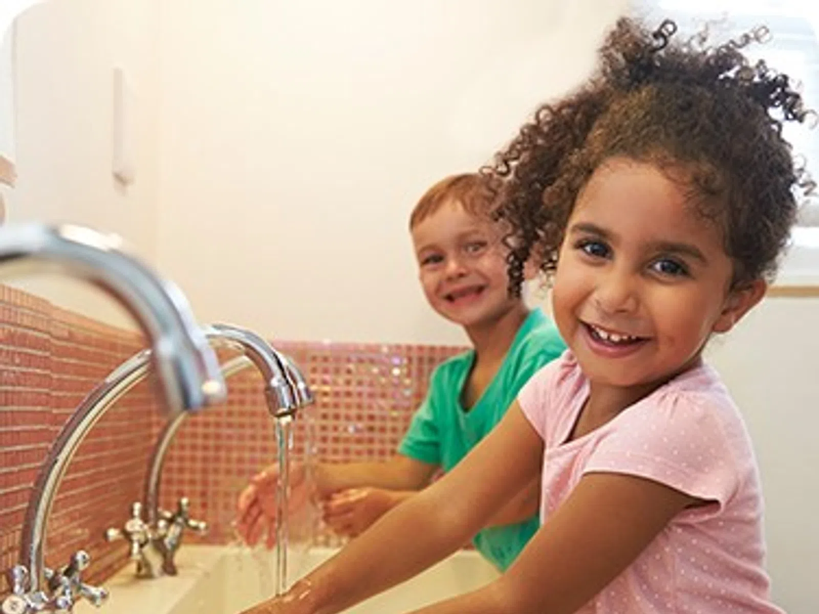 Two smiling children washing hands at a sink with running water in a bright bathroom.