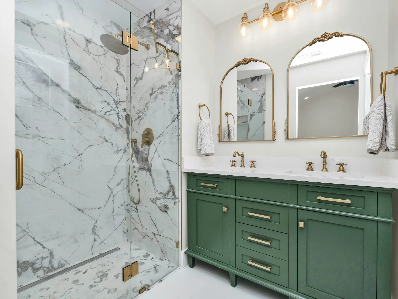 Modern bathroom with green vanity, white marble shower enclosure, gold fixtures, and double mirrors with lighting.