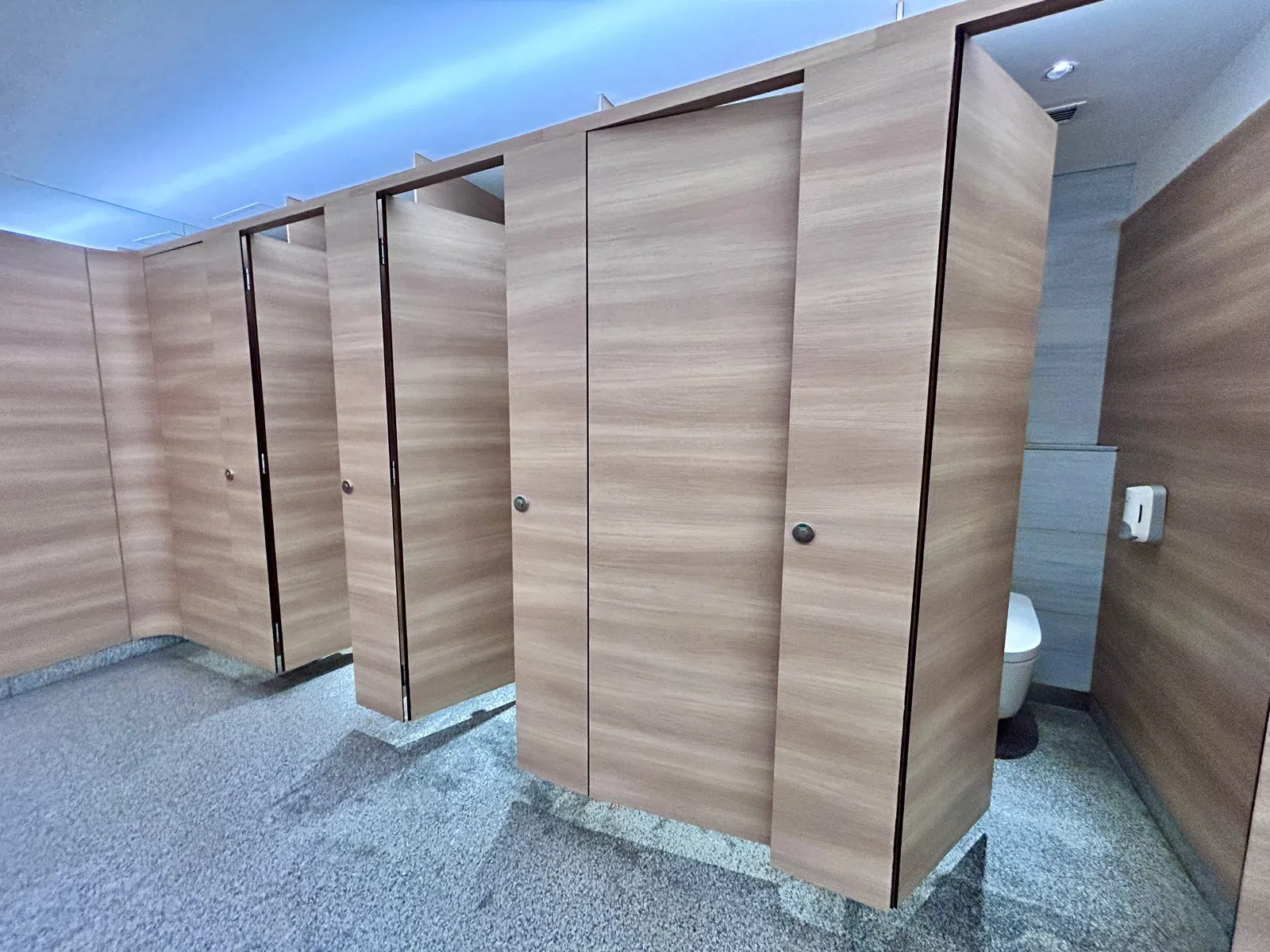 Modern public restroom with wood-toned stall doors and gray textured flooring under bright lighting.