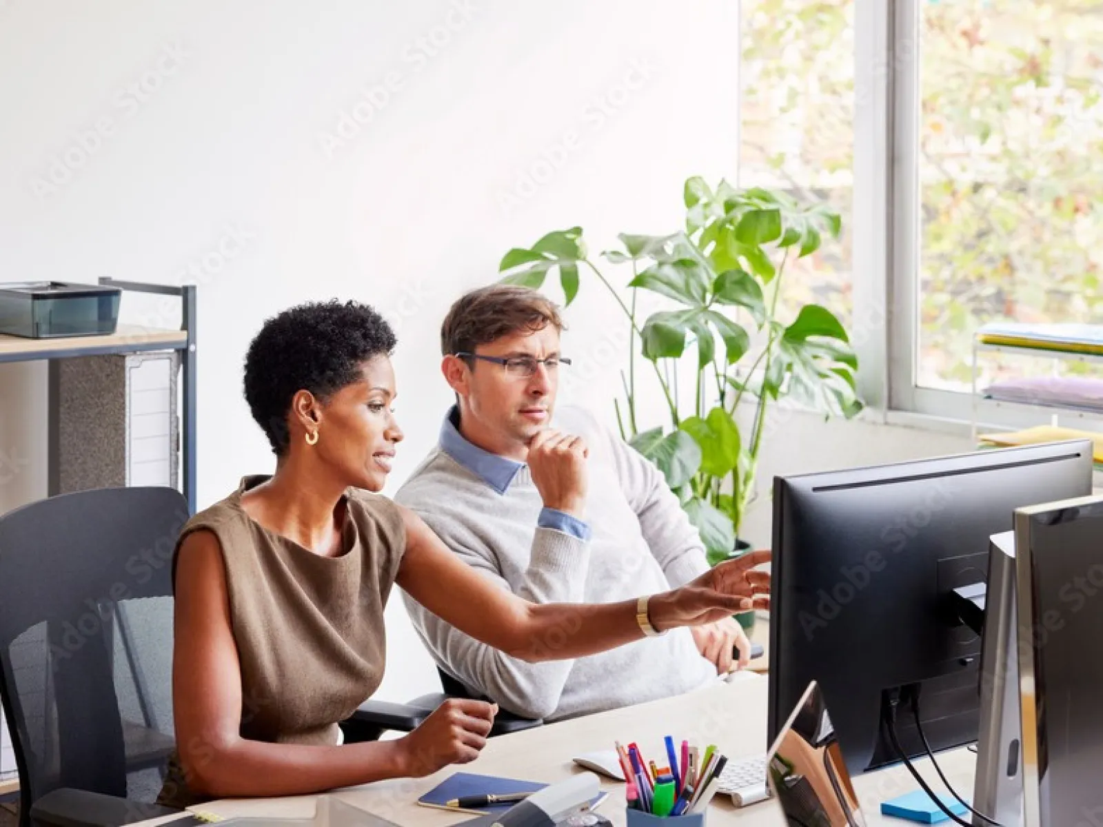 Two colleagues collaborate at a desk, focused on a computer screen in a bright modern office with plants.