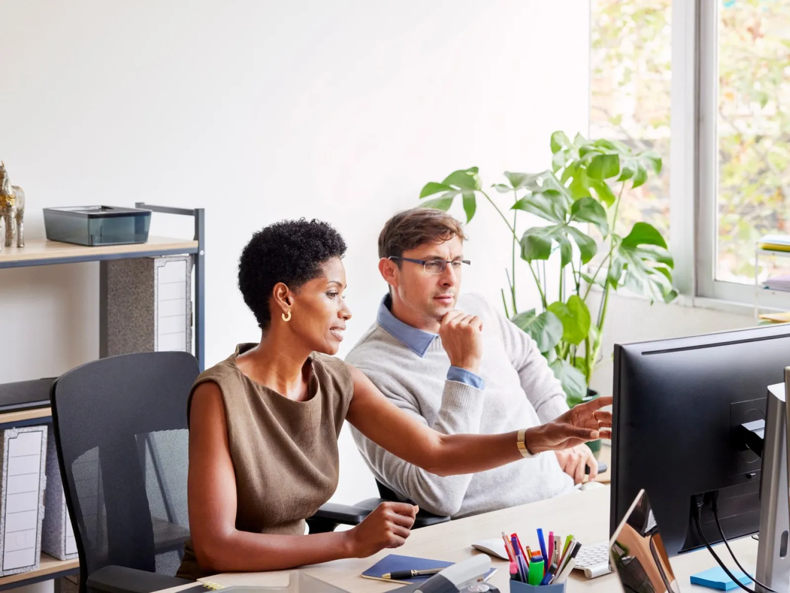 Two professionals collaborating and discussing work at a computer in a bright modern office with plants.