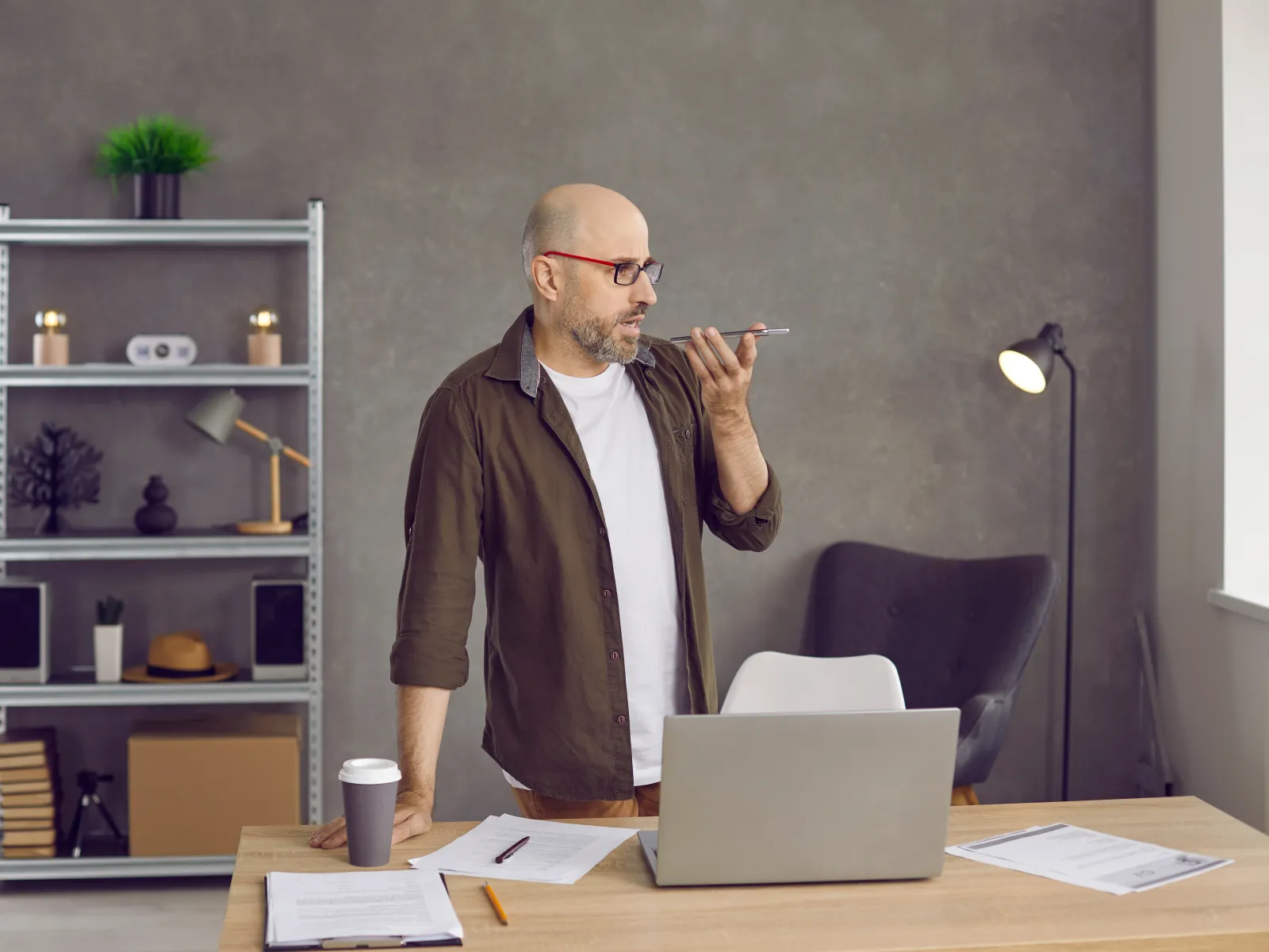 Bald man with glasses using voice command on smartphone in a modern home office setting with laptop and documents.