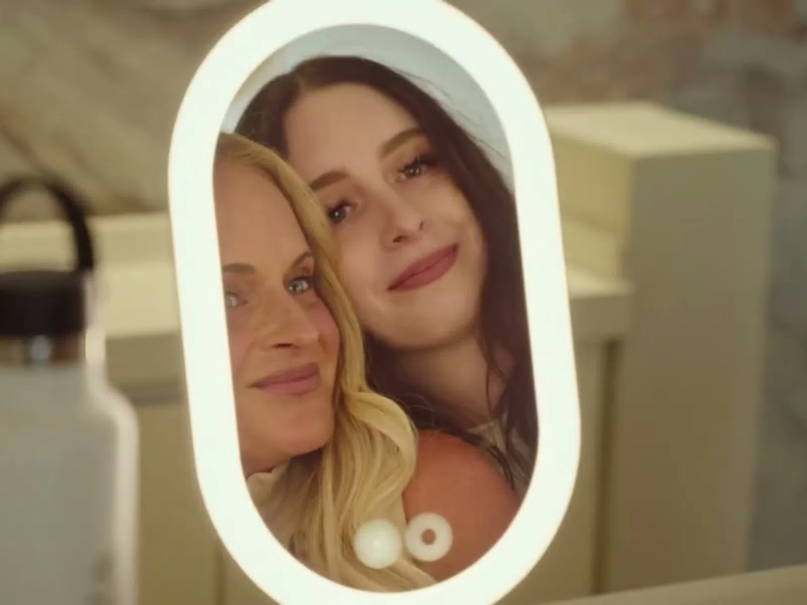 Two women smiling and taking a selfie reflected in an oval LED mirror with a water bottle in the foreground.