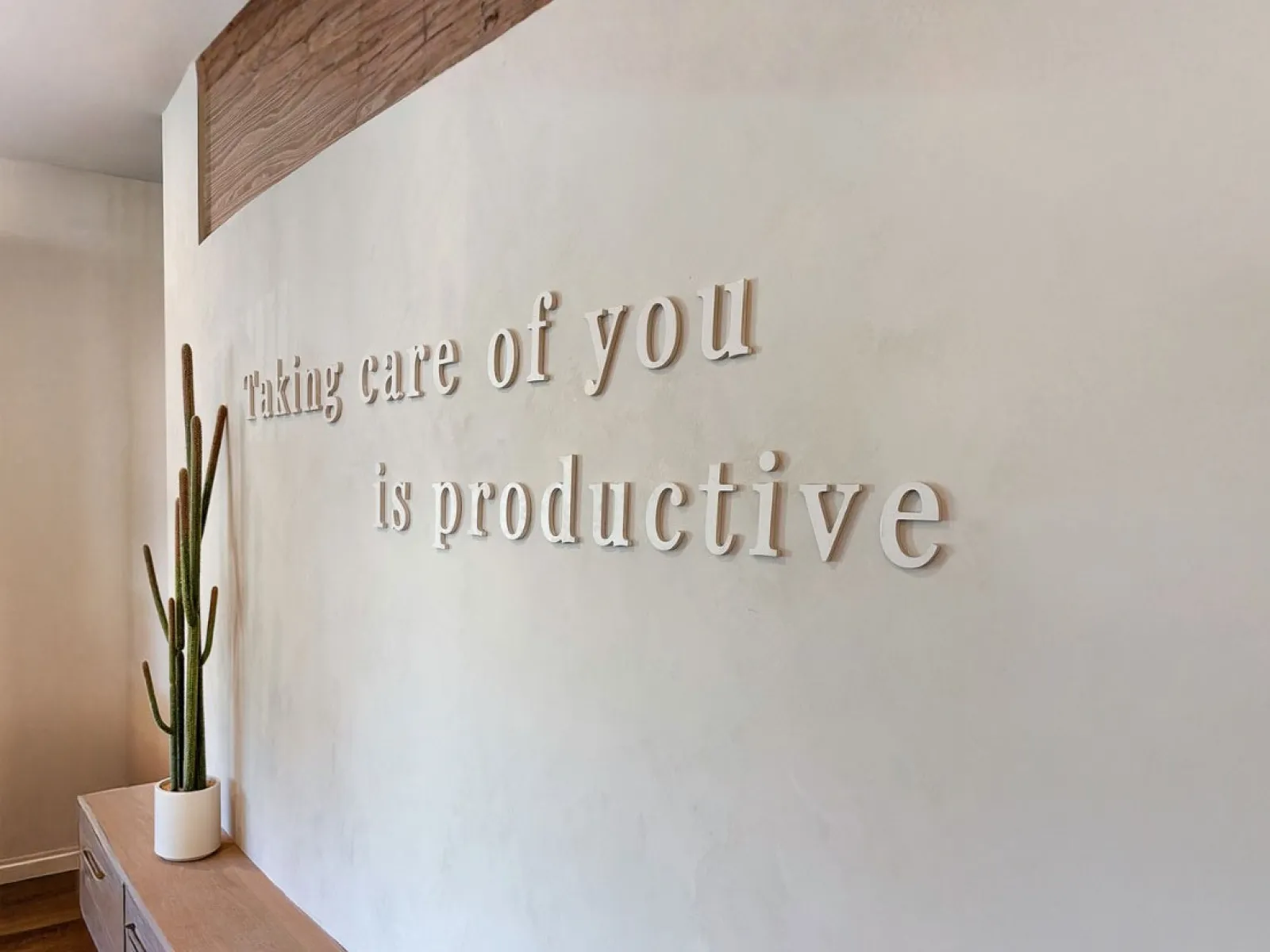 Minimalist wall with the phrase 'Taking care of you is productive' and a potted cactus on wooden furniture.