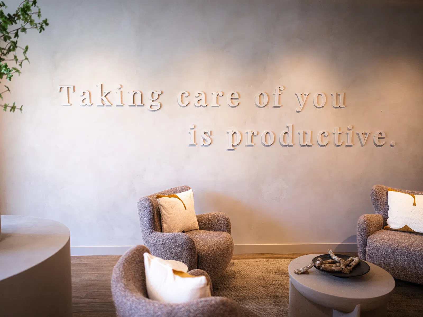 Cozy seating area with armchairs and pillows beneath a wall sign reading Taking care of you is productive.