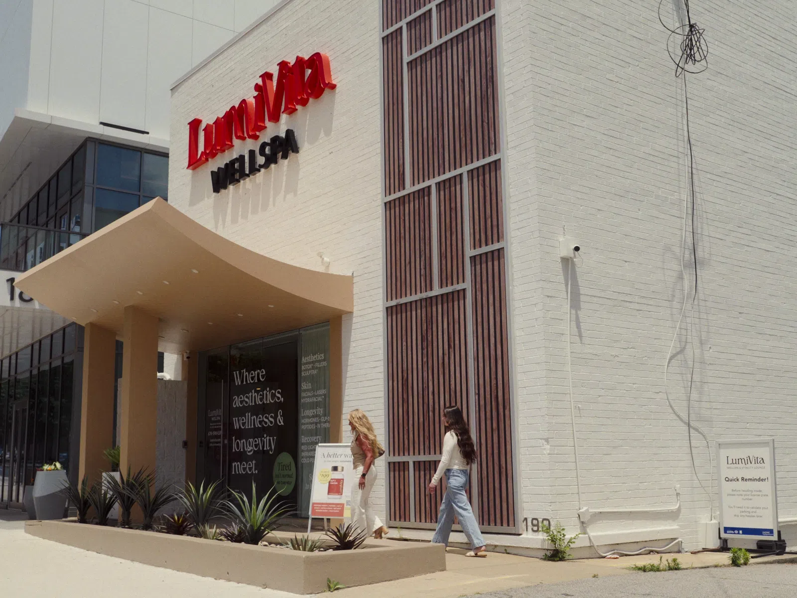 Two women walking past the white exterior of Lumivita Wellness & Spa with red signage on the building.