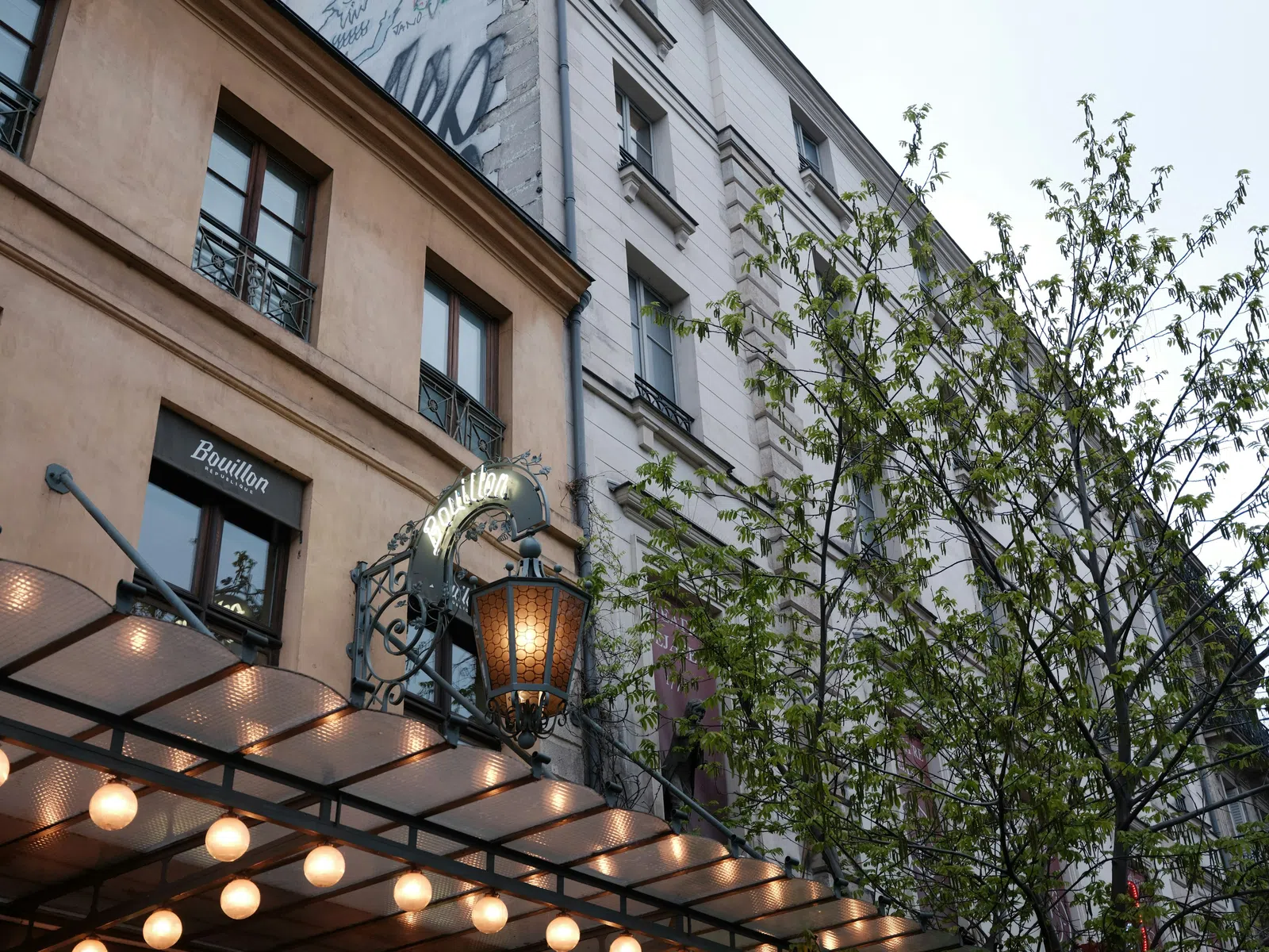 Exterior of Bouillon restaurant with glowing marquee lights and tree in front of Parisian buildings at dusk