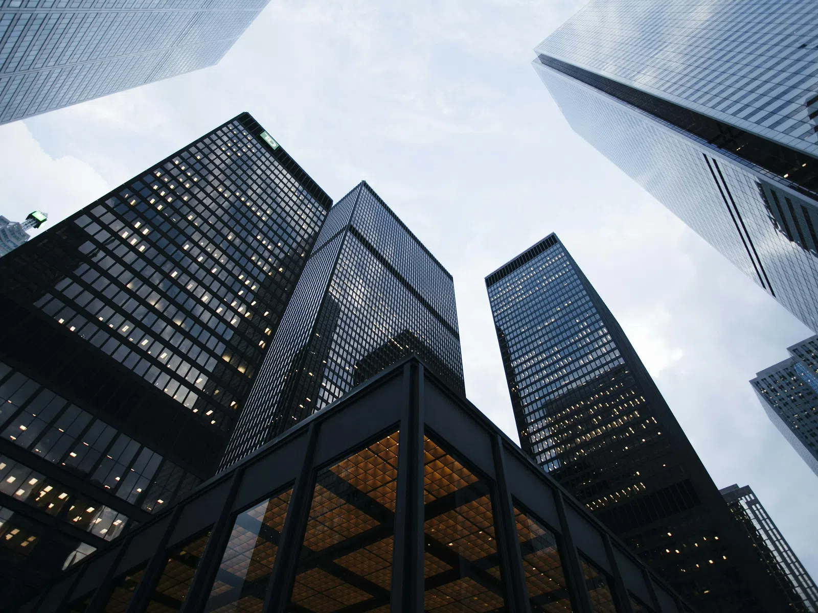 View looking up at modern glass skyscrapers with lit windows against a cloudy sky in a city center.