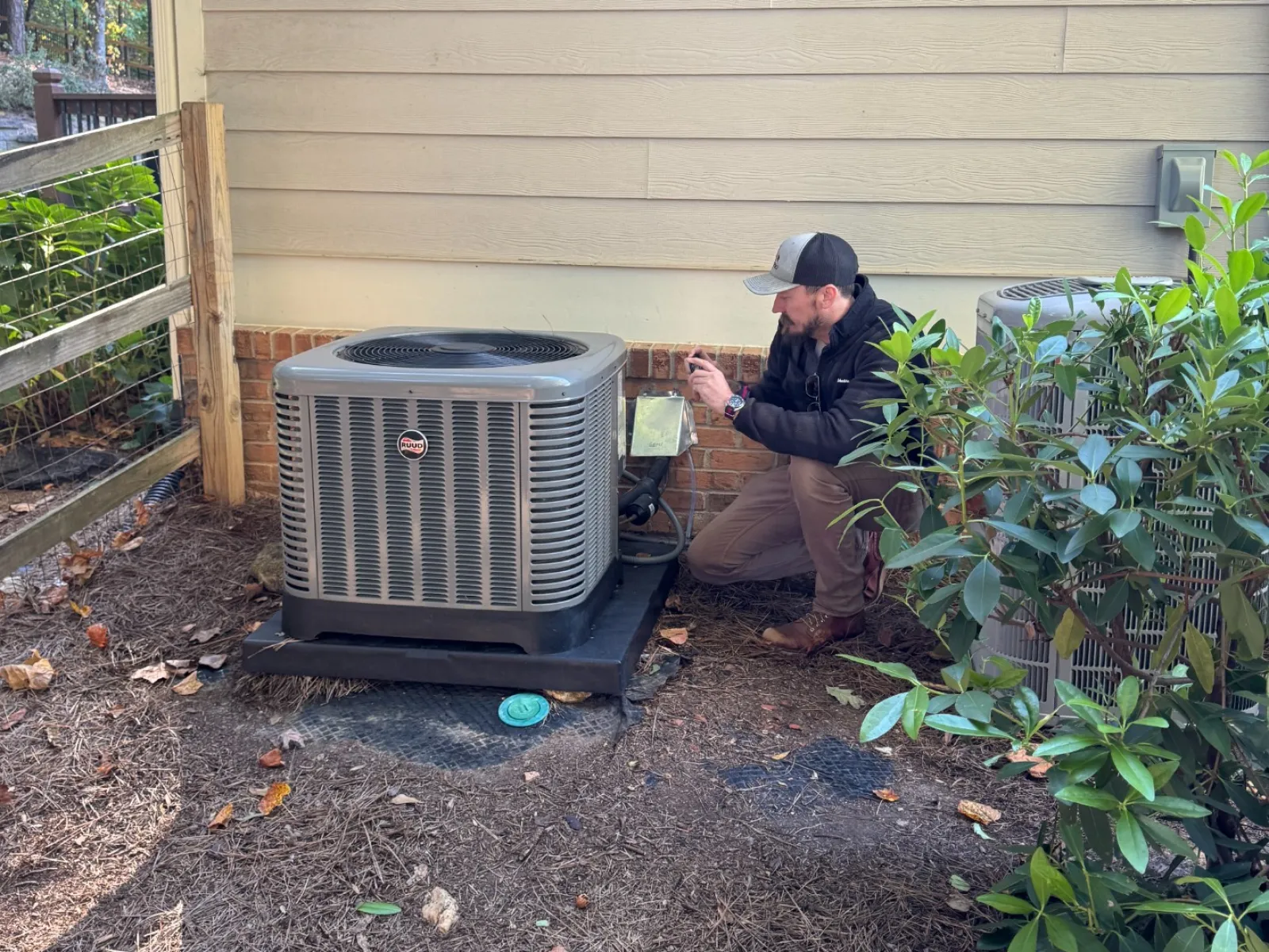 Technician inspecting an outdoor HVAC unit next to a beige house with surrounding plants and mulch.