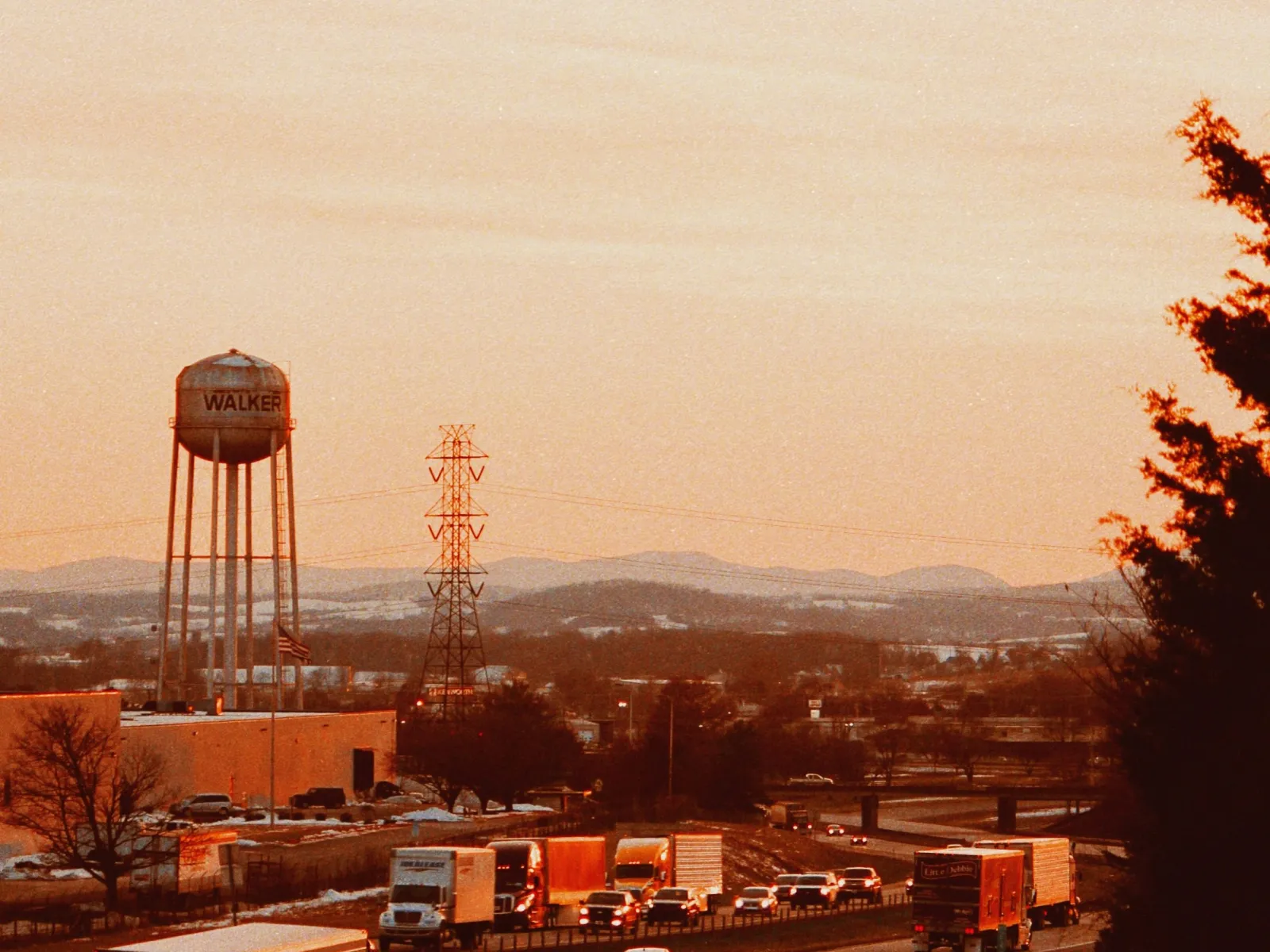 Trucks and cars driving on highway near water tower and power lines at sunset in rural area.