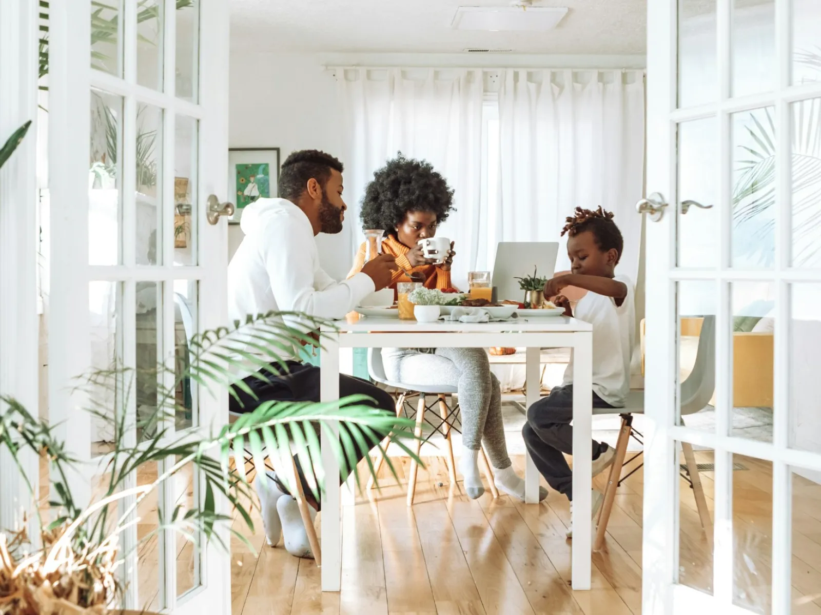 Family of three enjoying breakfast together at a white dining table in a bright, plant-filled room.