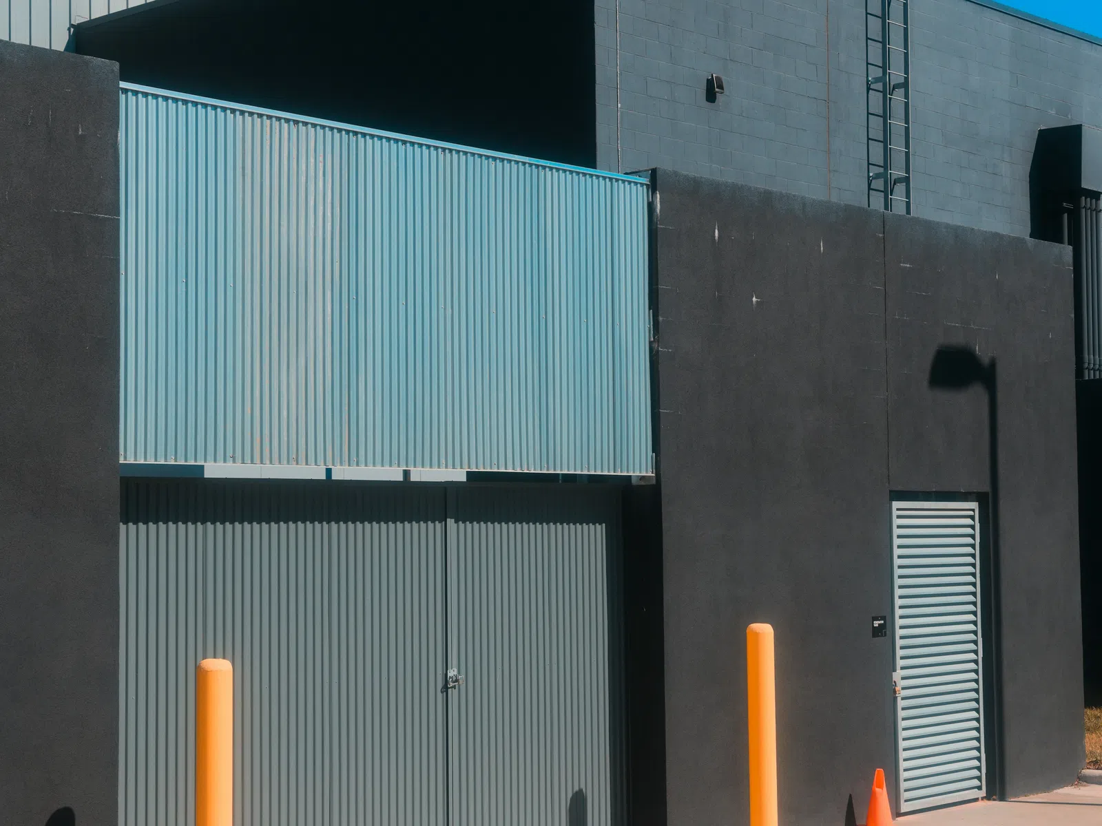 Industrial building facade with corrugated metal doors, bright yellow bollards, and clear blue sky overhead