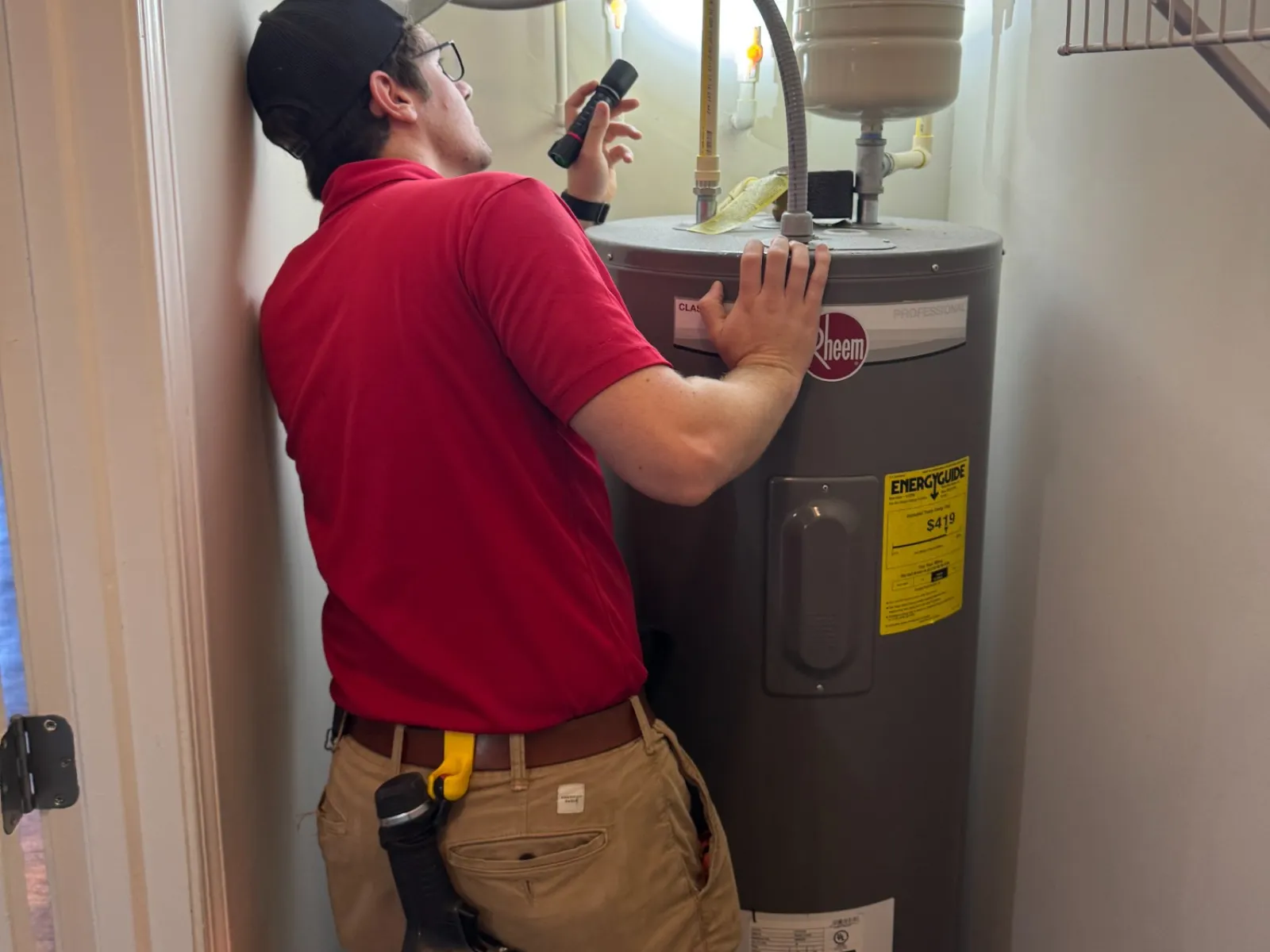 Technician inspecting and repairing a residential water heater with a flashlight in a utility closet
