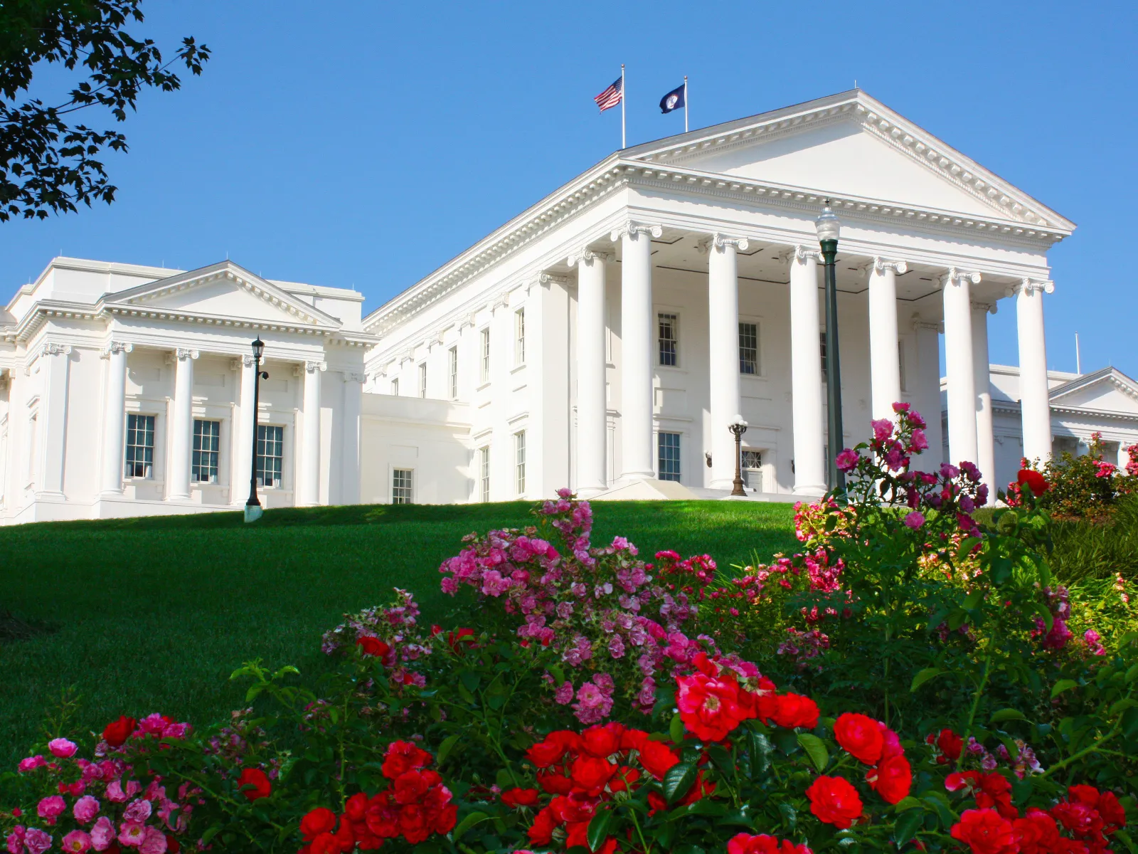 a white building with columns and a garden of flowers in front of it with Virginia State Capitol in the background