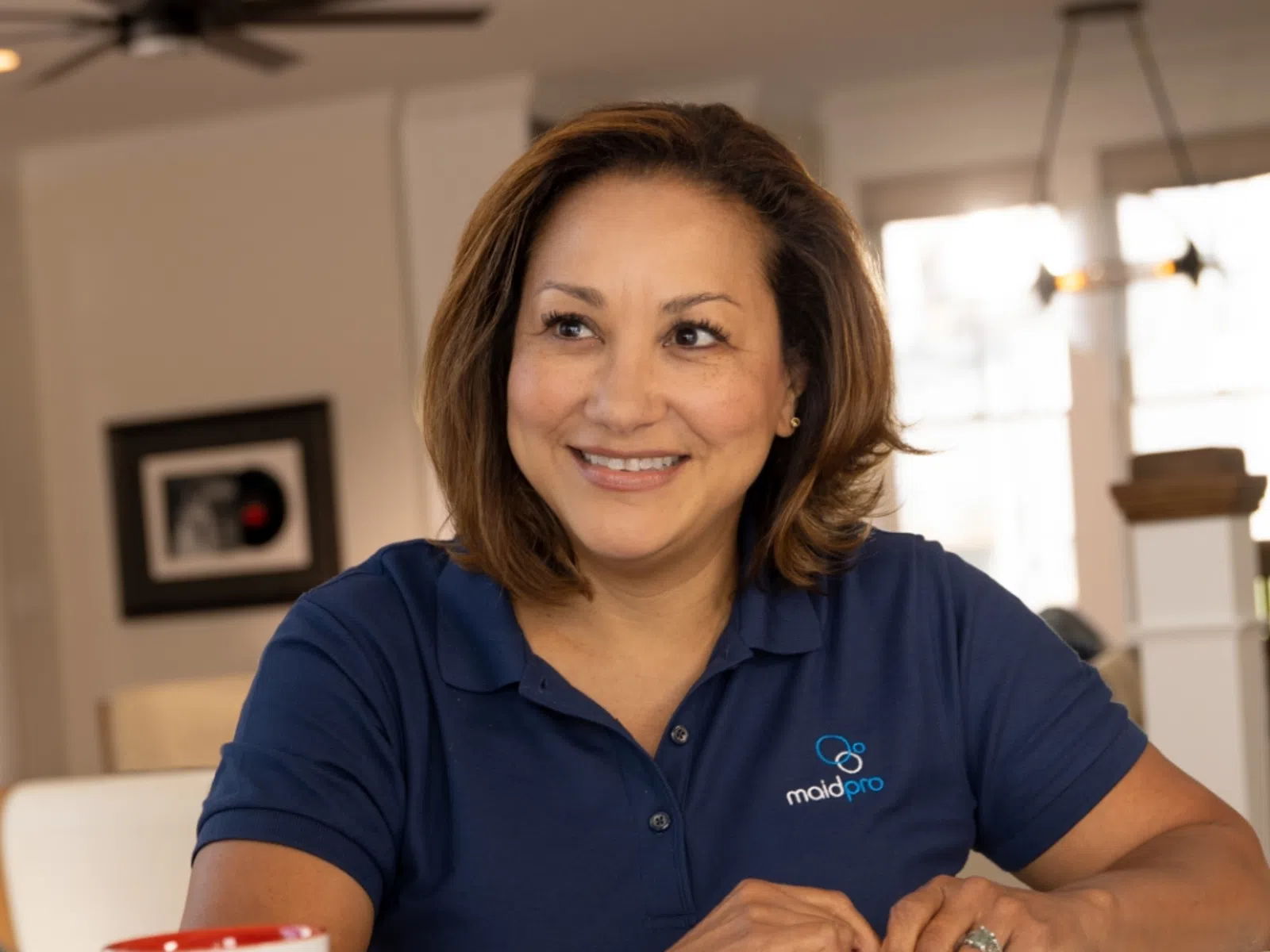 Smiling woman in a MaidPro polo shirt chatting with another woman at a table in a cozy home setting