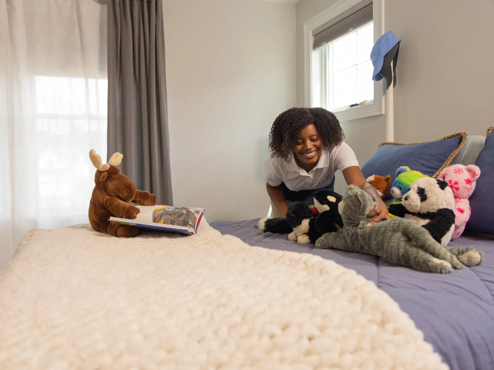 Smiling woman arranging various stuffed animals on a neatly made bed in a bright bedroom.