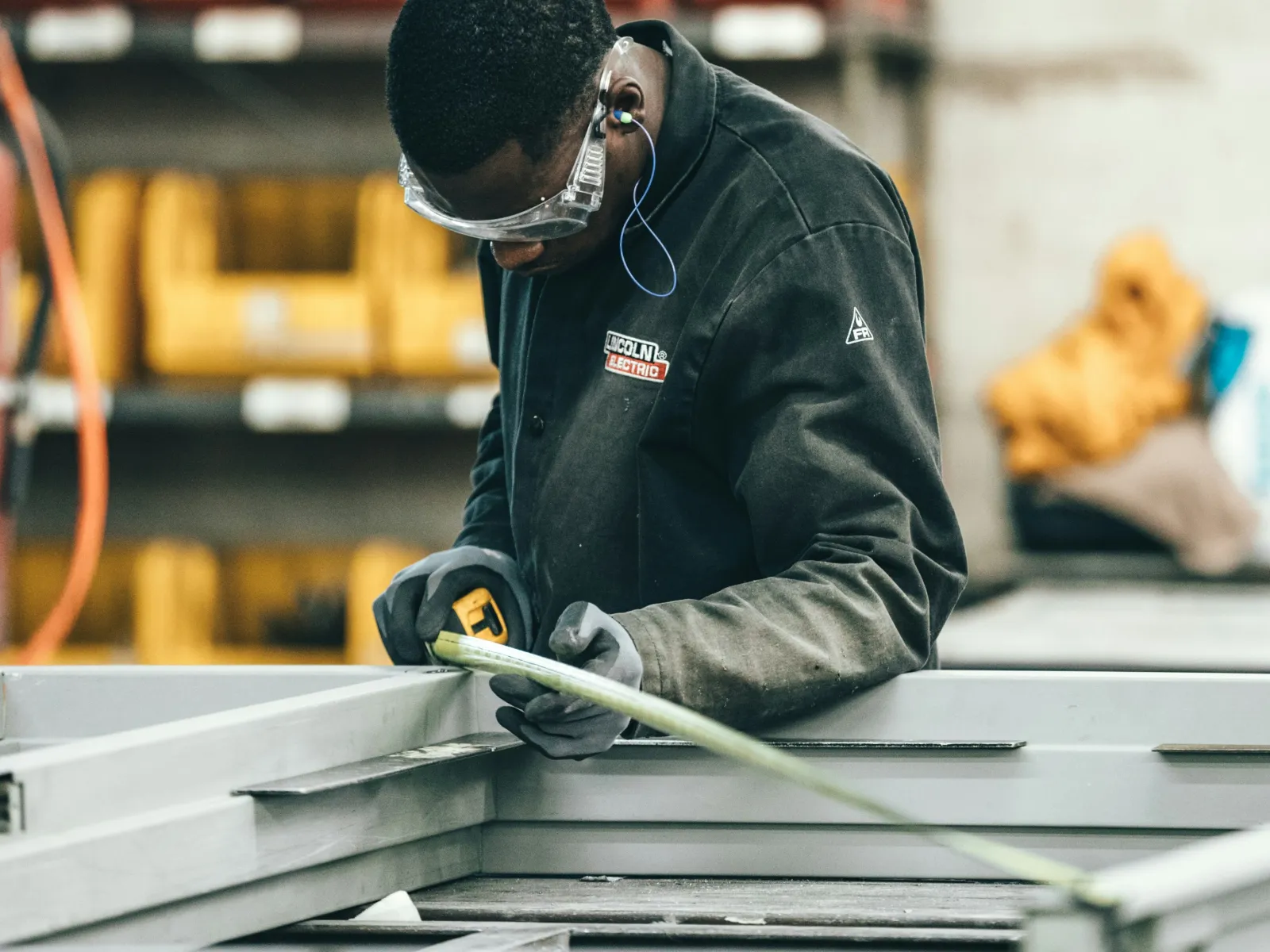 Worker in protective gear measuring metal frame with tape in industrial workshop setting.