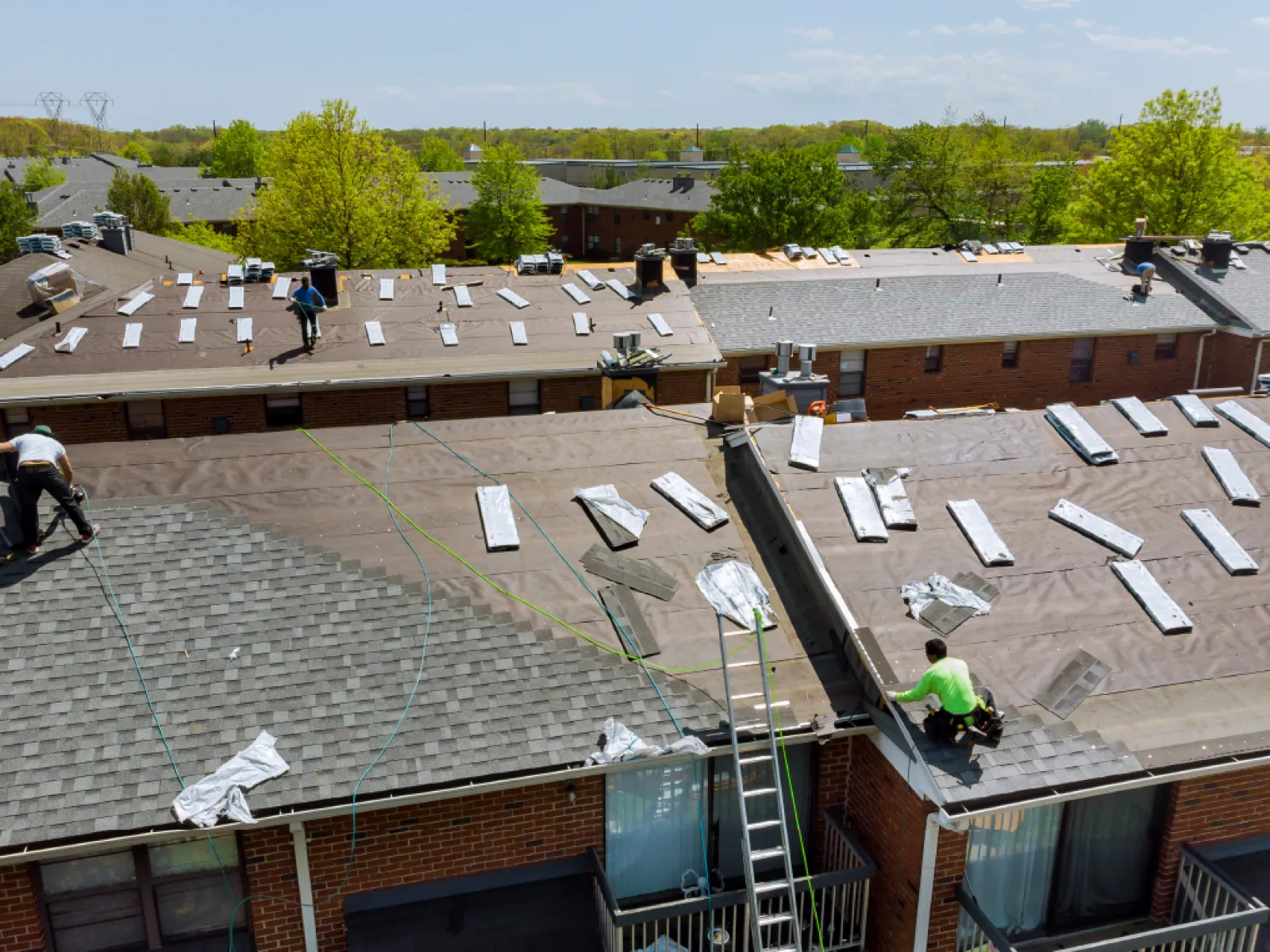 Workers installing roofs on apartment buildings under a clear blue sky surrounded by greenery.