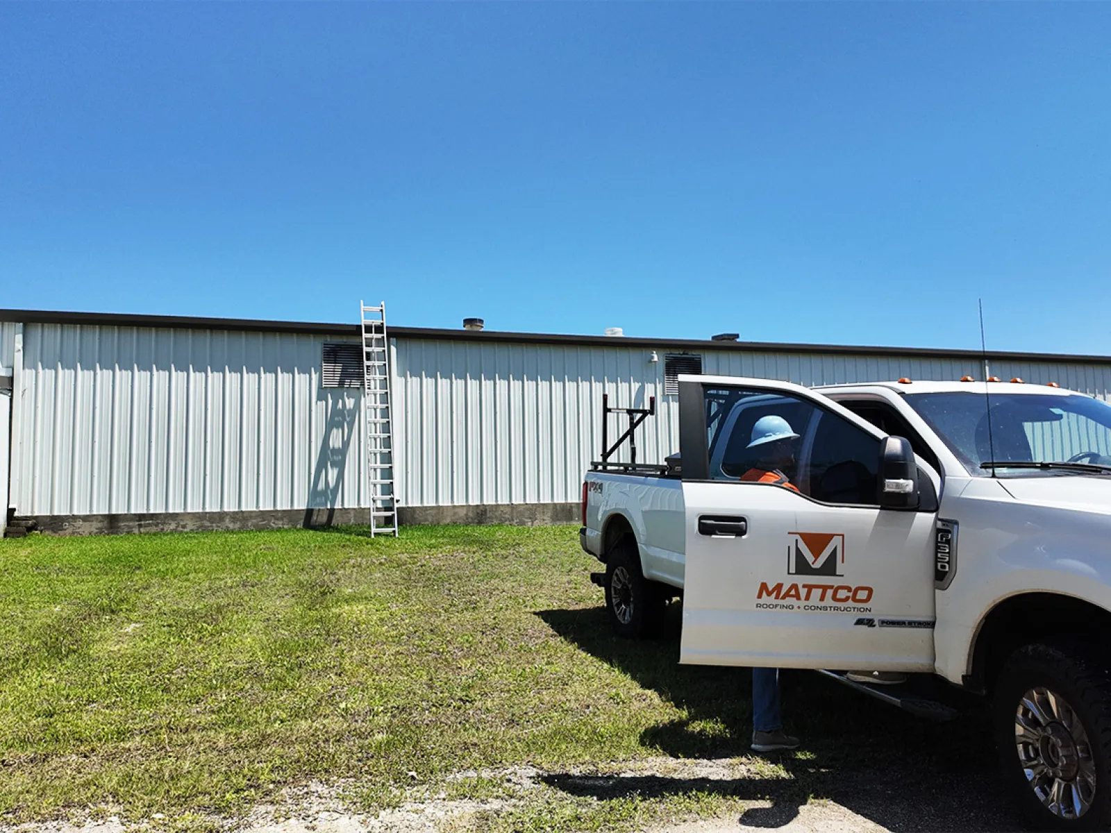 Mattco Roofing and Construction branded work truck parked on site at a commercial metal building in Texas with a ladder leaning against the exterior wall and a crew member in an orange safety vest and hard hat preparing for a rooftop inspection visit.