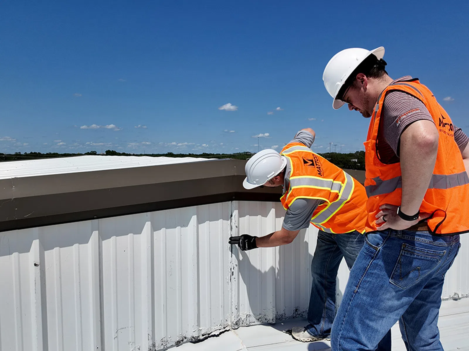 Two Mattco roofing technicians in orange safety vests inspecting a commercial metal roof edge and parapet wall on a clear day in Texas. One technician is standing and examining the flashing detail while the other crouches to assess the base of the wall. Mattco branding is visible on the standing technician's vest.