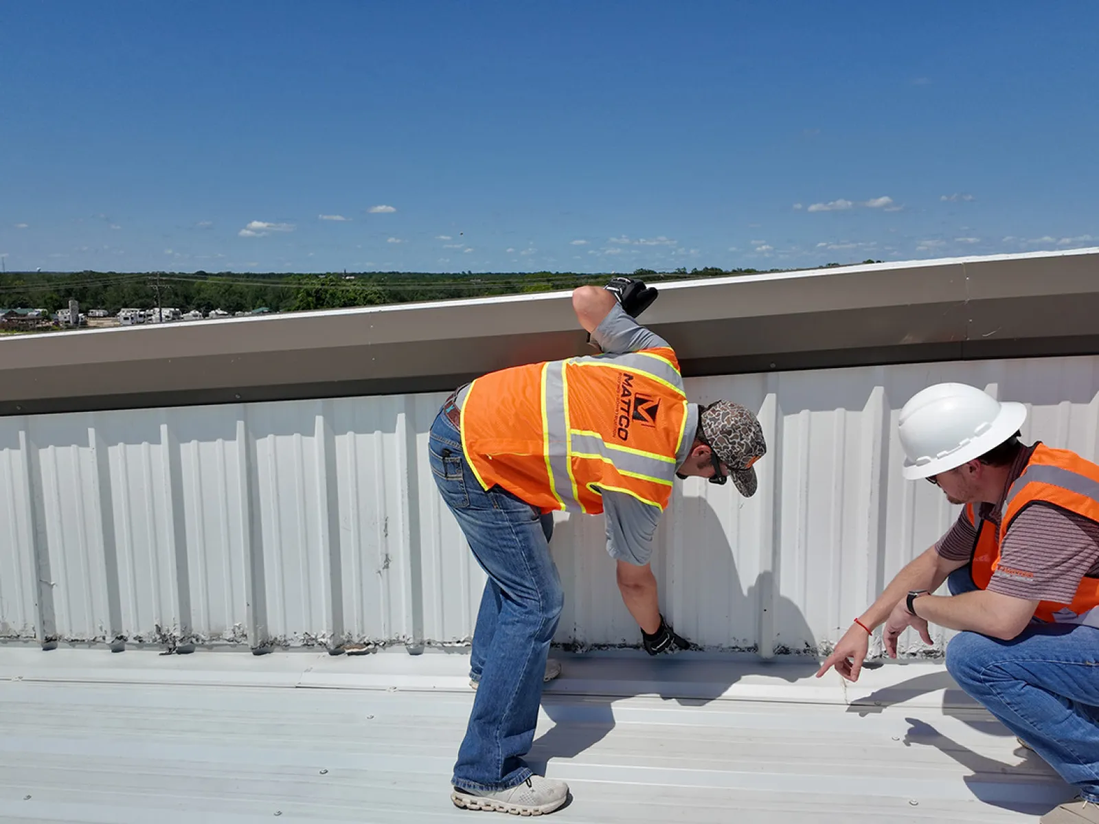Two Mattco roofing technicians in orange safety vests inspecting a commercial metal roof edge and parapet wall on a clear day in Texas. One technician is standing and examining the flashing detail while the other crouches to assess the base of the wall. Mattco branding is visible on the standing technician's vest.