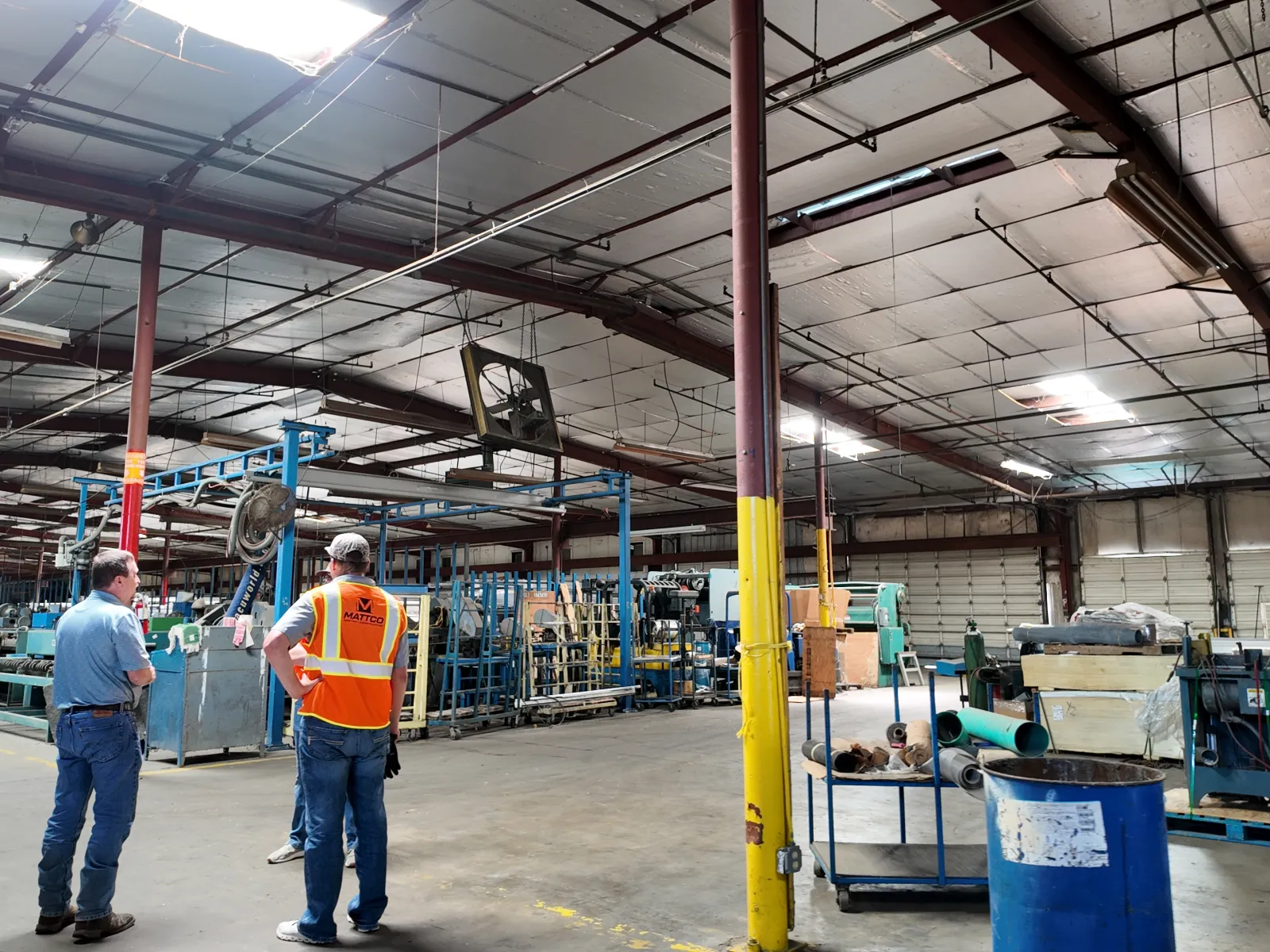 Two workers in a large industrial warehouse with machinery and equipment under high ceilings and bright lighting.