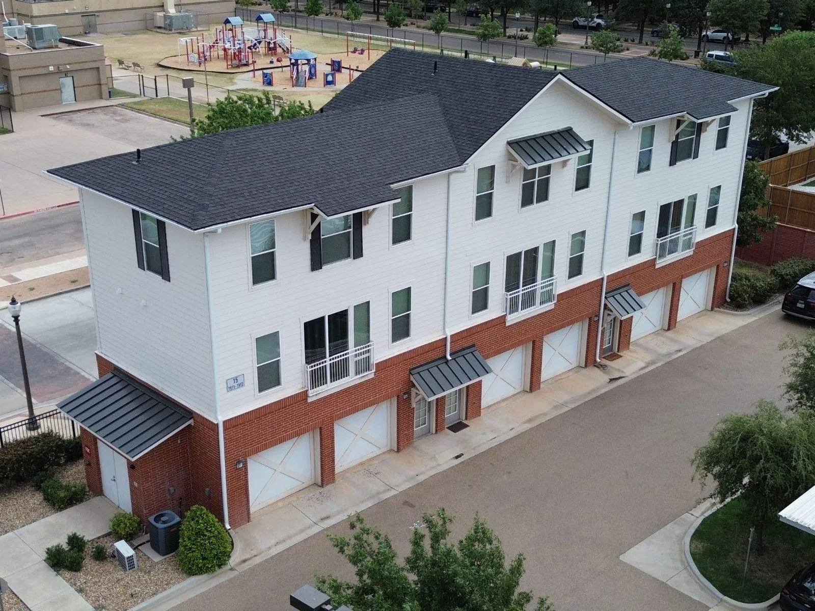 Modern three-story townhouse with white siding and brick exterior in urban residential area with playground nearby