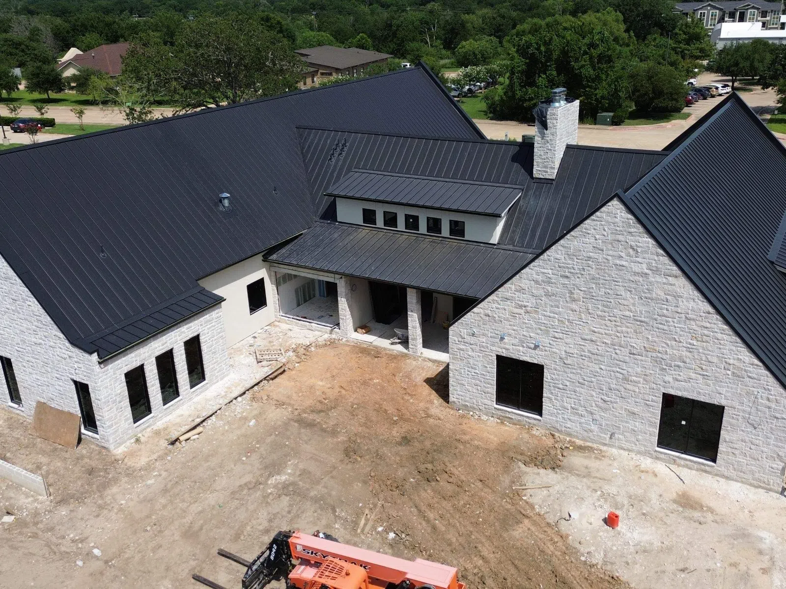 A large modern house with white stone walls and a black metal roof under construction in a suburban area.