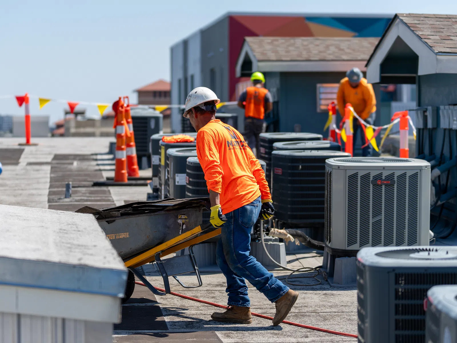 Construction workers in safety gear working on rooftop HVAC units with orange safety cones and equipment